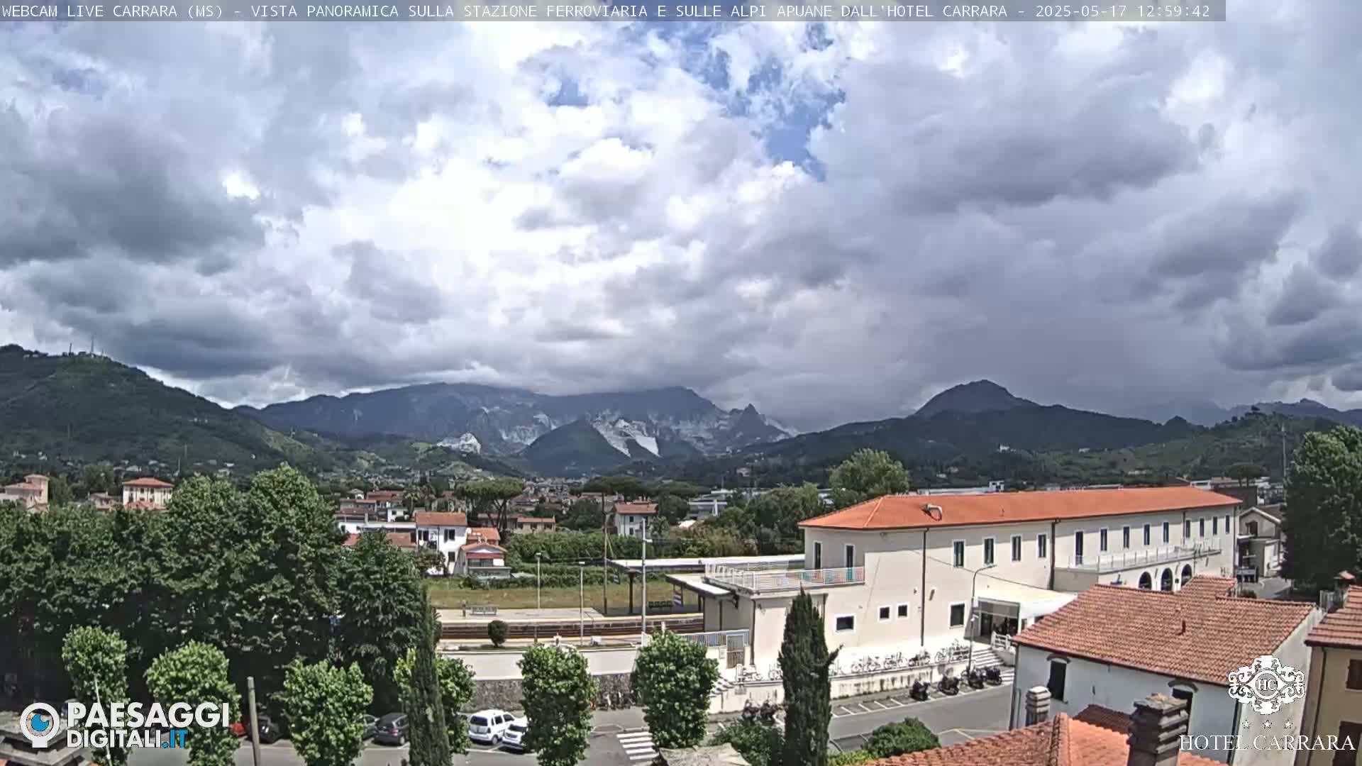 A partly cloudy day reveals a view of a town nestled in a valley between mountains, with a long building, possibly a hotel or train station, in the foreground.