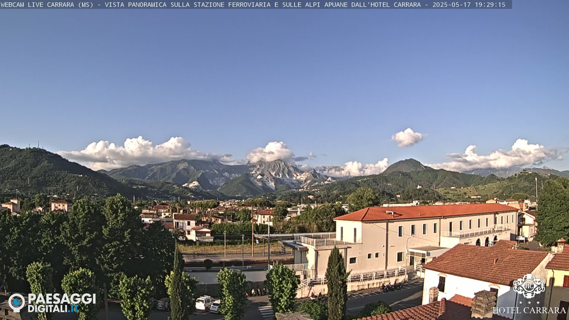 A partly cloudy day reveals a panoramic view of a town nestled in a valley, surrounded by lush green hills and mountains under a clear blue sky.