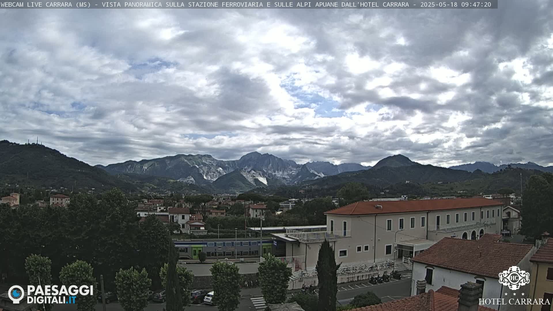 A partly cloudy day reveals a panoramic view of a town nestled in a valley, with mountains in the background and a train visible near a large building.