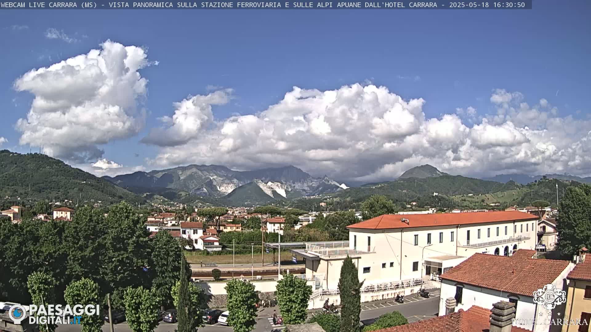 A partly cloudy day reveals a town nestled in a valley, with mountains in the background and a long, low building in the foreground.