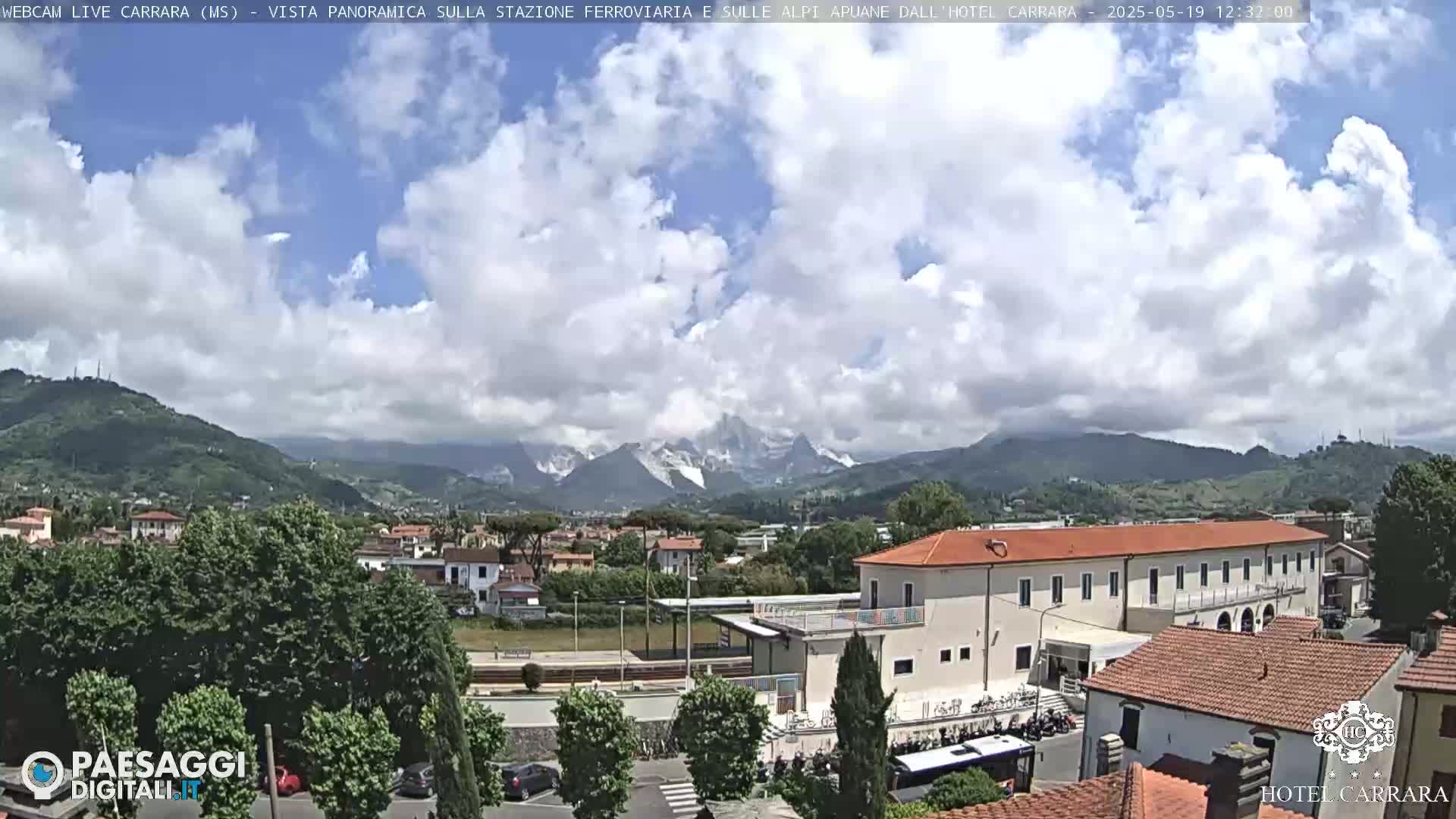 A partly cloudy day reveals a view of a town nestled in a valley, with snow-capped mountains in the distance and a large building near the foreground.