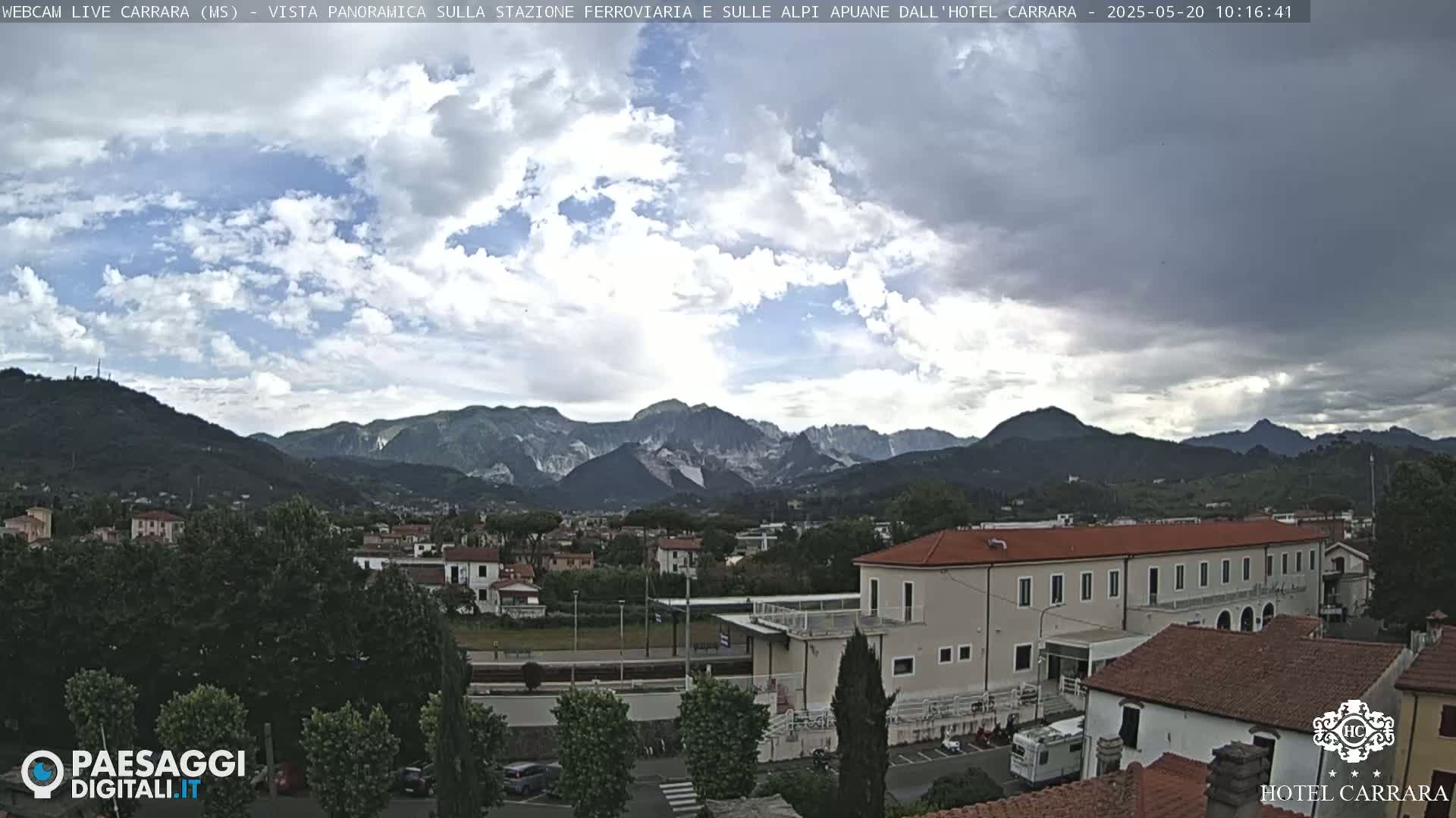 A partly cloudy day reveals a panoramic view of a town nestled in a valley, with mountains in the background and a building prominently in the foreground.