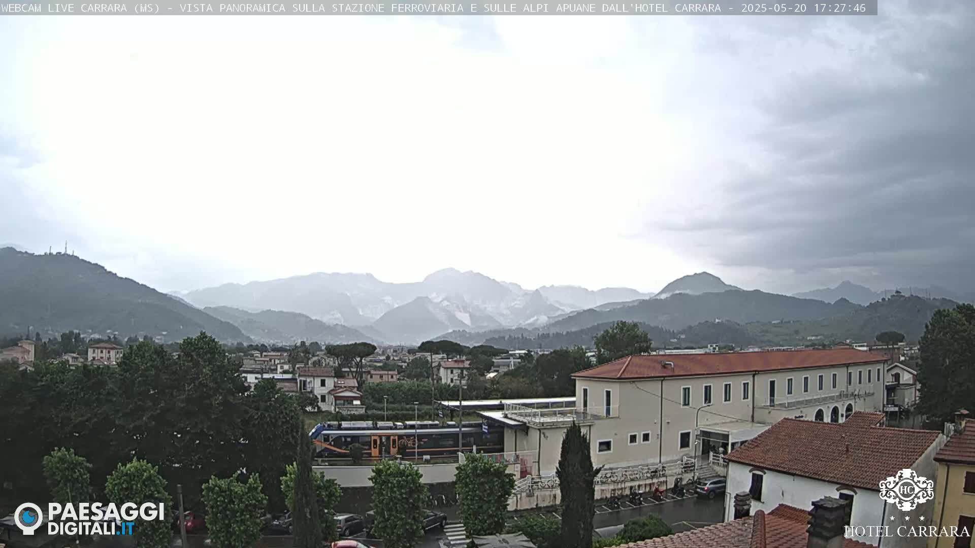 A train is visible near a long building, with mountains in the background under an overcast sky.