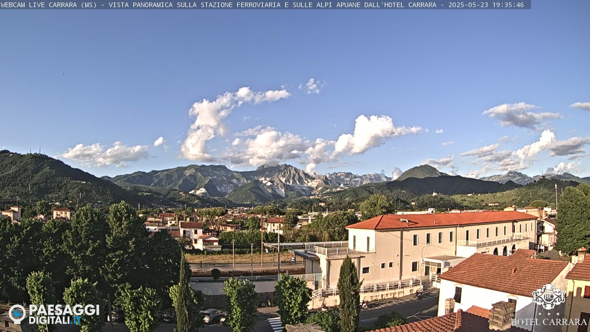 A partly sunny view of a town nestled in a valley, with mountains in the background and a large building in the foreground.