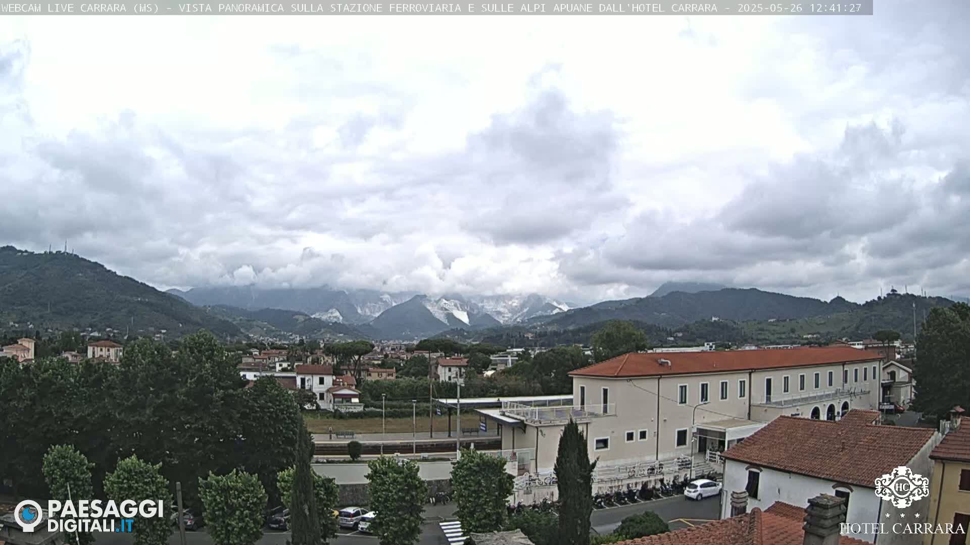 A partly cloudy day reveals a panoramic view of a town nestled in a valley, with mountains in the background, one of which has a snowy peak.