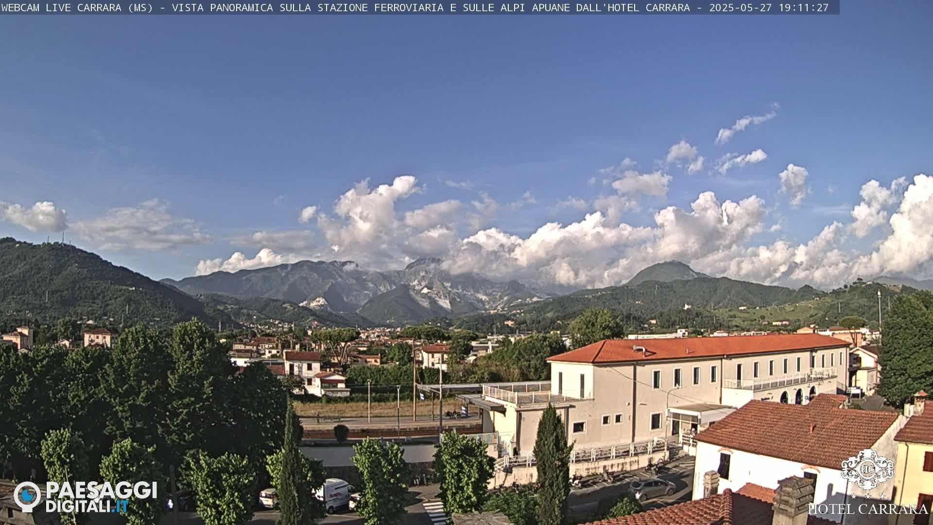 A partly cloudy day shows a view of a town nestled in a valley, with mountains in the background and a large building in the foreground.