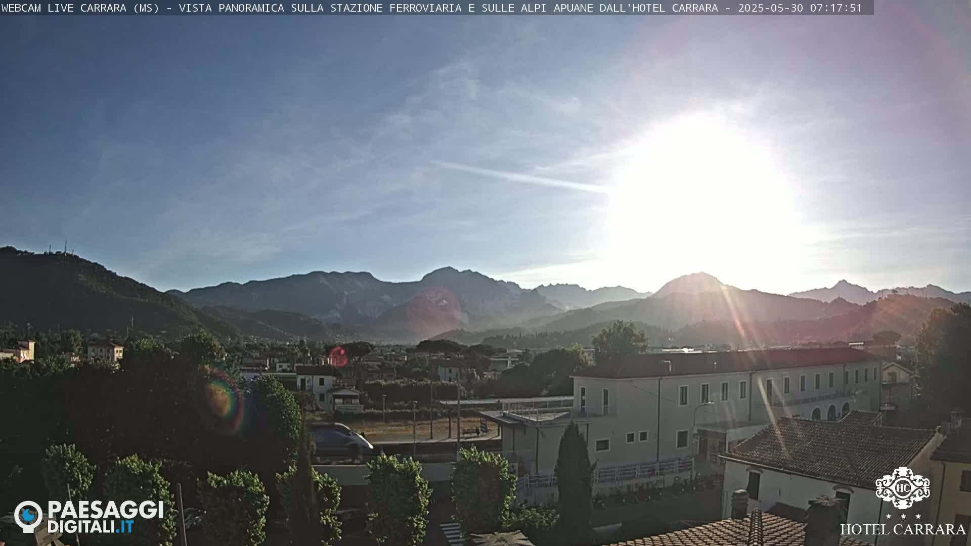 A sunny, bright-looking view of a town nestled in a valley, with mountains in the background and a train partially visible near buildings.
