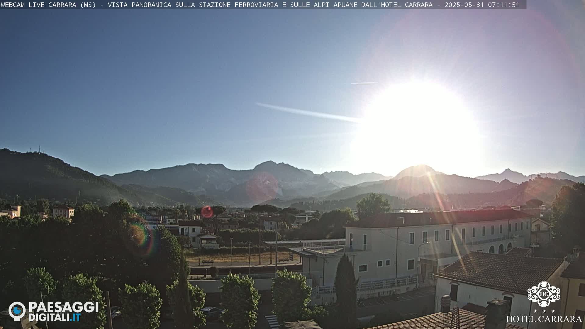 A sunny morning view shows a town nestled in a valley, with mountains in the background and a bright sun casting rays.