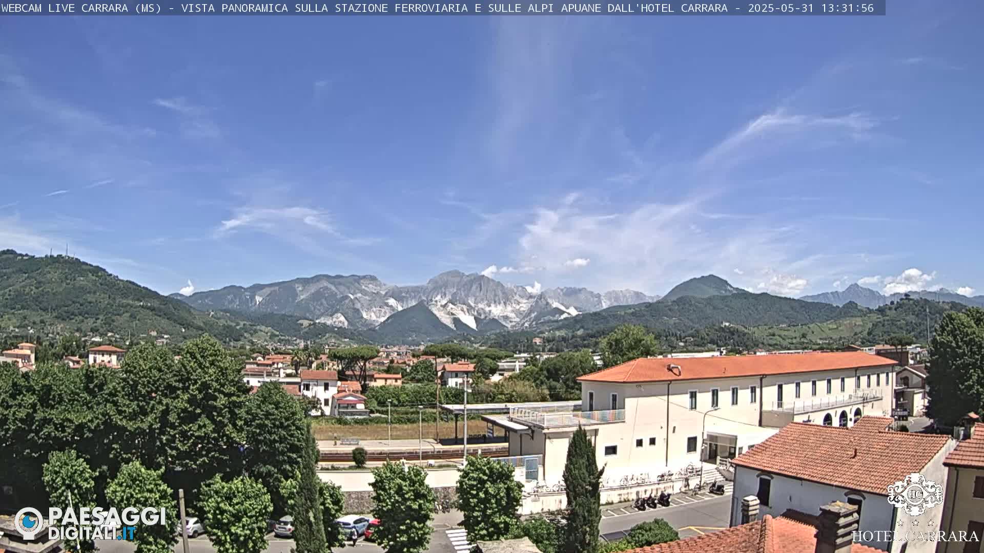 A sunny day reveals a panoramic view of a town nestled in a valley, with a long building near the foreground and a mountain range in the background.