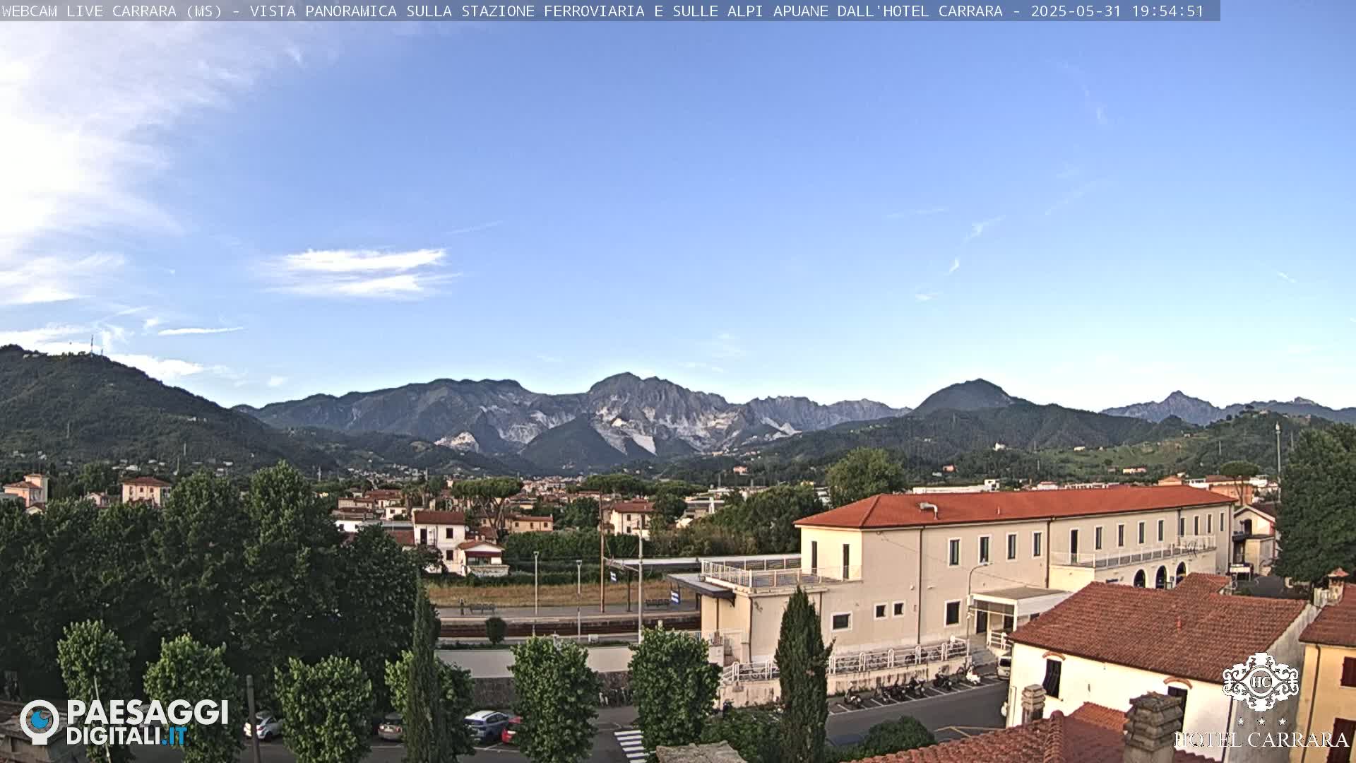 A panoramic view of a town nestled in a valley, with mountains in the background under a mostly clear sky.