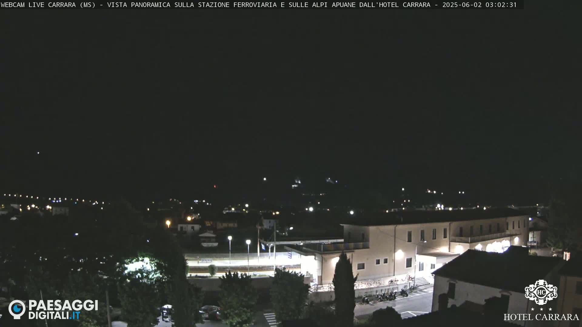 A nighttime long shot of a town's outskirts, showing a train station and a large building, under a clear, dark sky.
