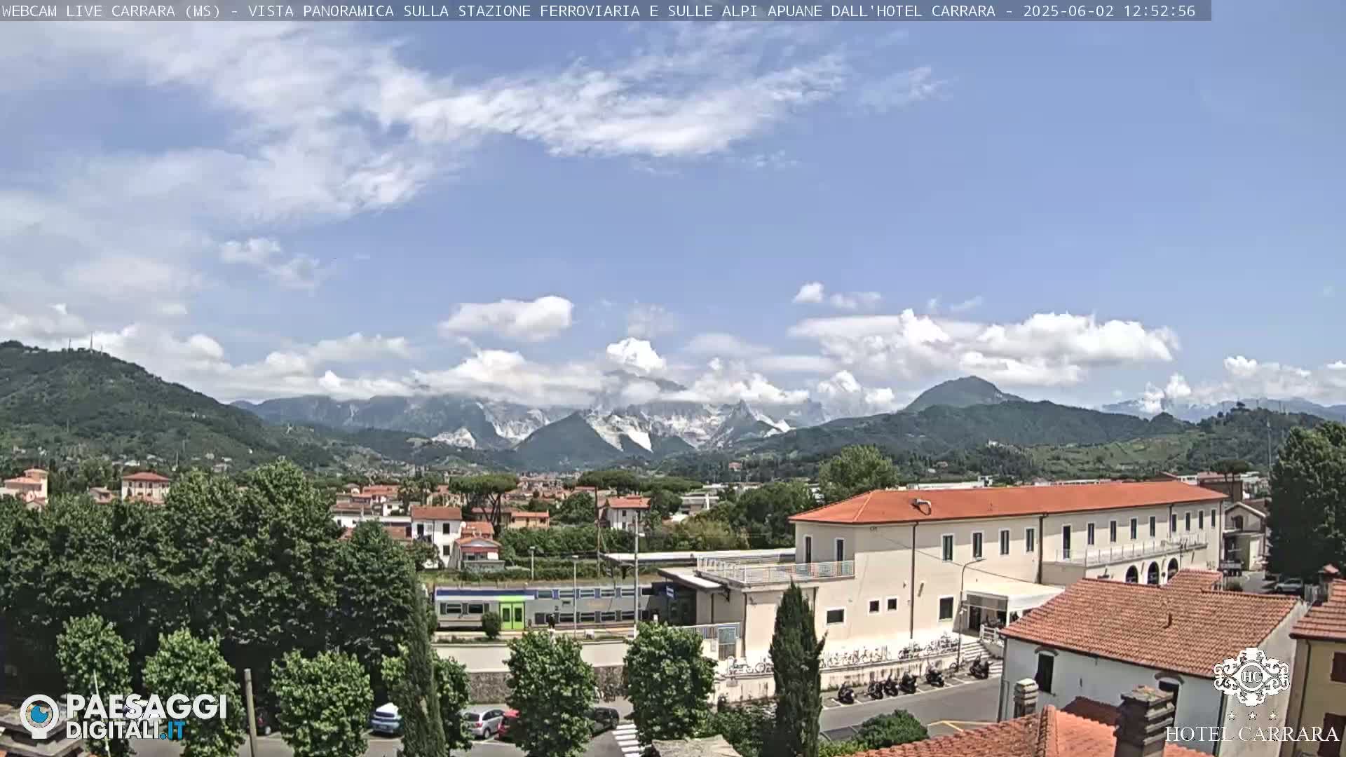 A partly cloudy day reveals a train station and surrounding buildings nestled in a valley, with mountains in the background, some showing snow-capped peaks.