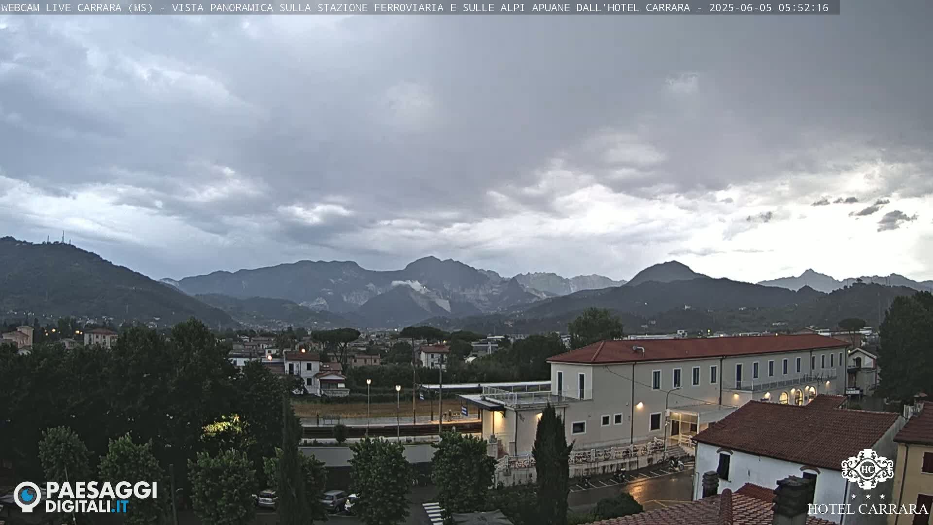 A long, low building is situated near a train station and surrounded by lush greenery, with a mountain range under an overcast sky.