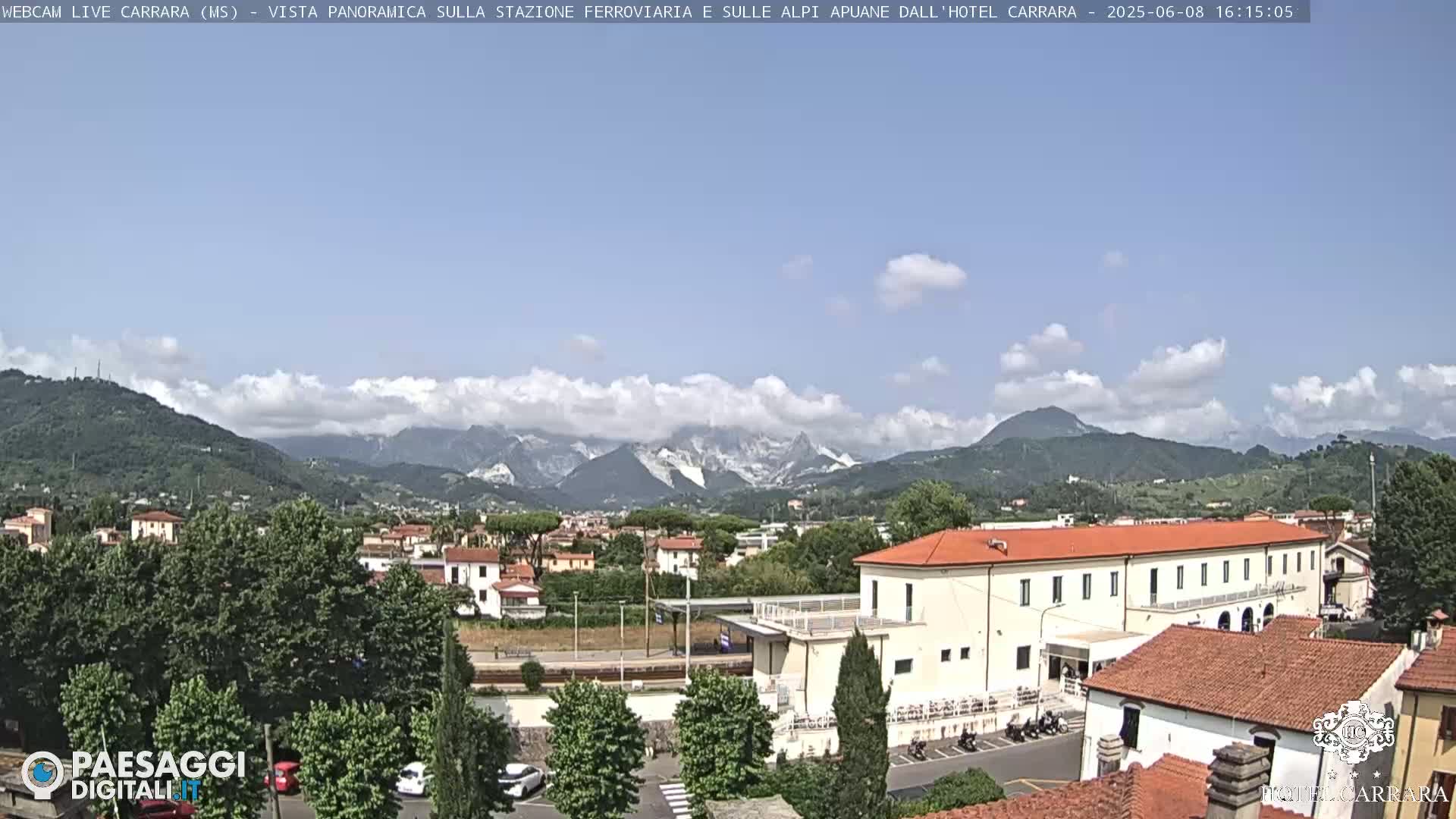A partly cloudy day reveals a panoramic view of a town nestled in a valley, with mountains in the background and a long, low building in the foreground.