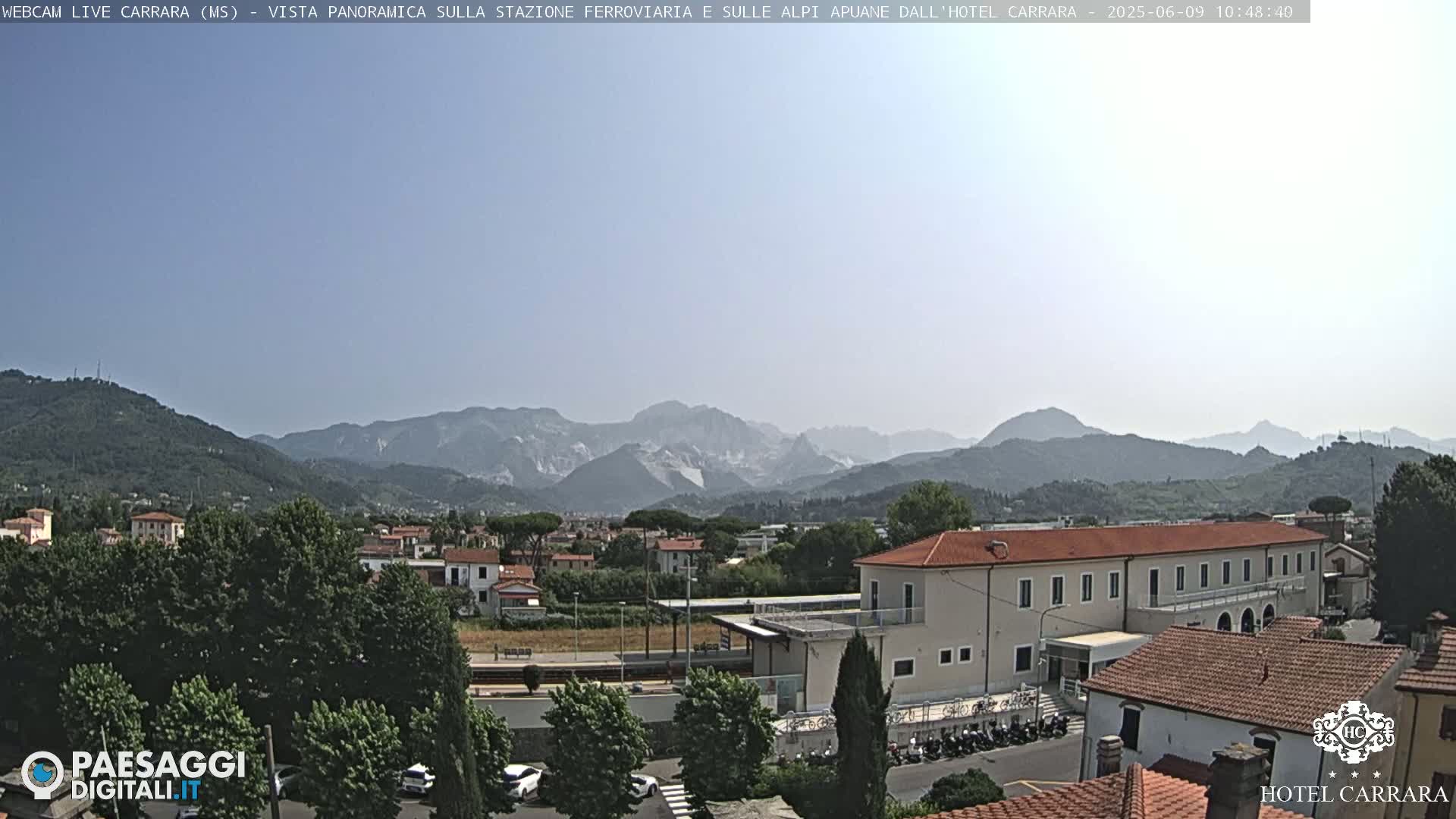A long, low building sits near a train station, with a mountain range visible in the distance under a hazy sky.