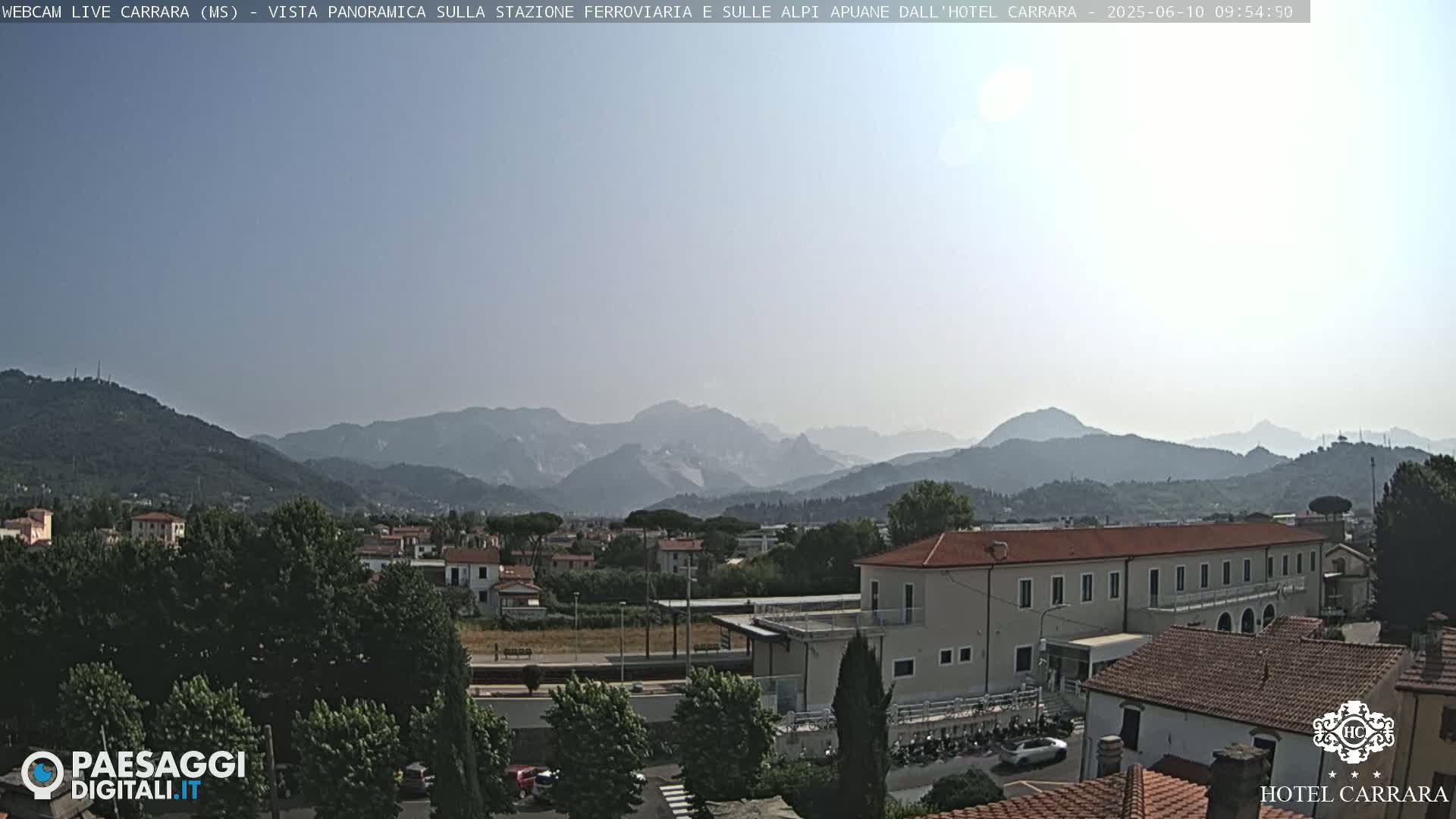 A hazy sunny day reveals a panoramic view of a town nestled among green hills and mountains in the distance.