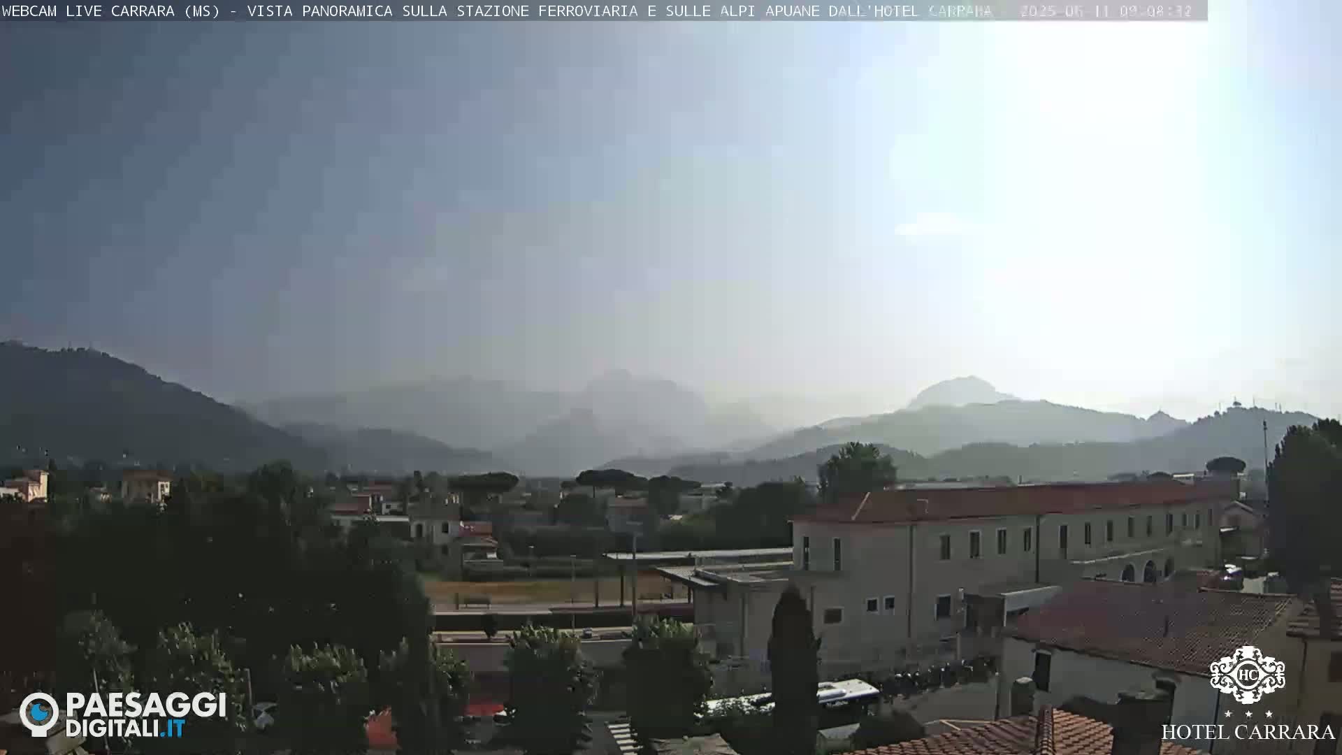 A hazy, sunny view of a town nestled between mountains and hills, with buildings and greenery visible.