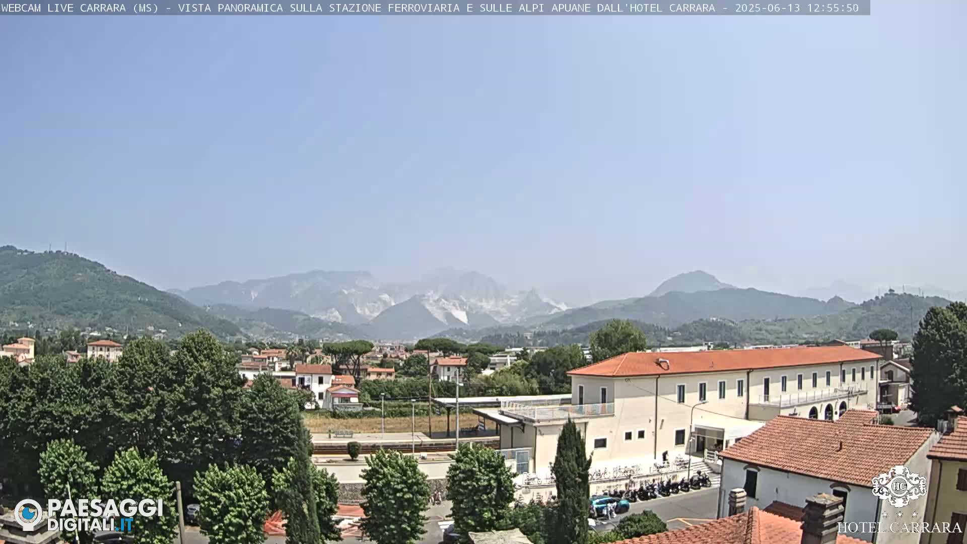 A long, low building is situated near a train station, with a mountainous landscape visible in the background under a clear, sunny sky.