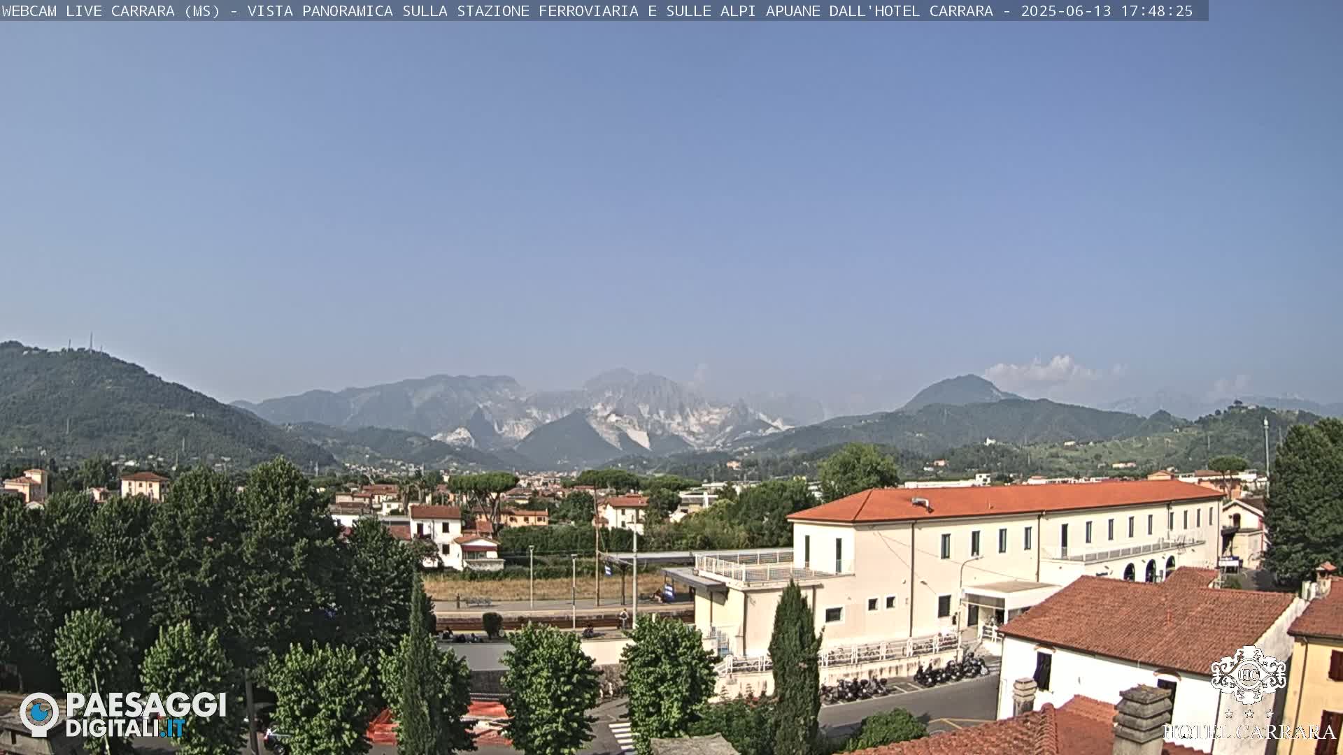 A long shot shows a clear, sunny day with mountains in the background and a town with buildings and trees in the foreground.