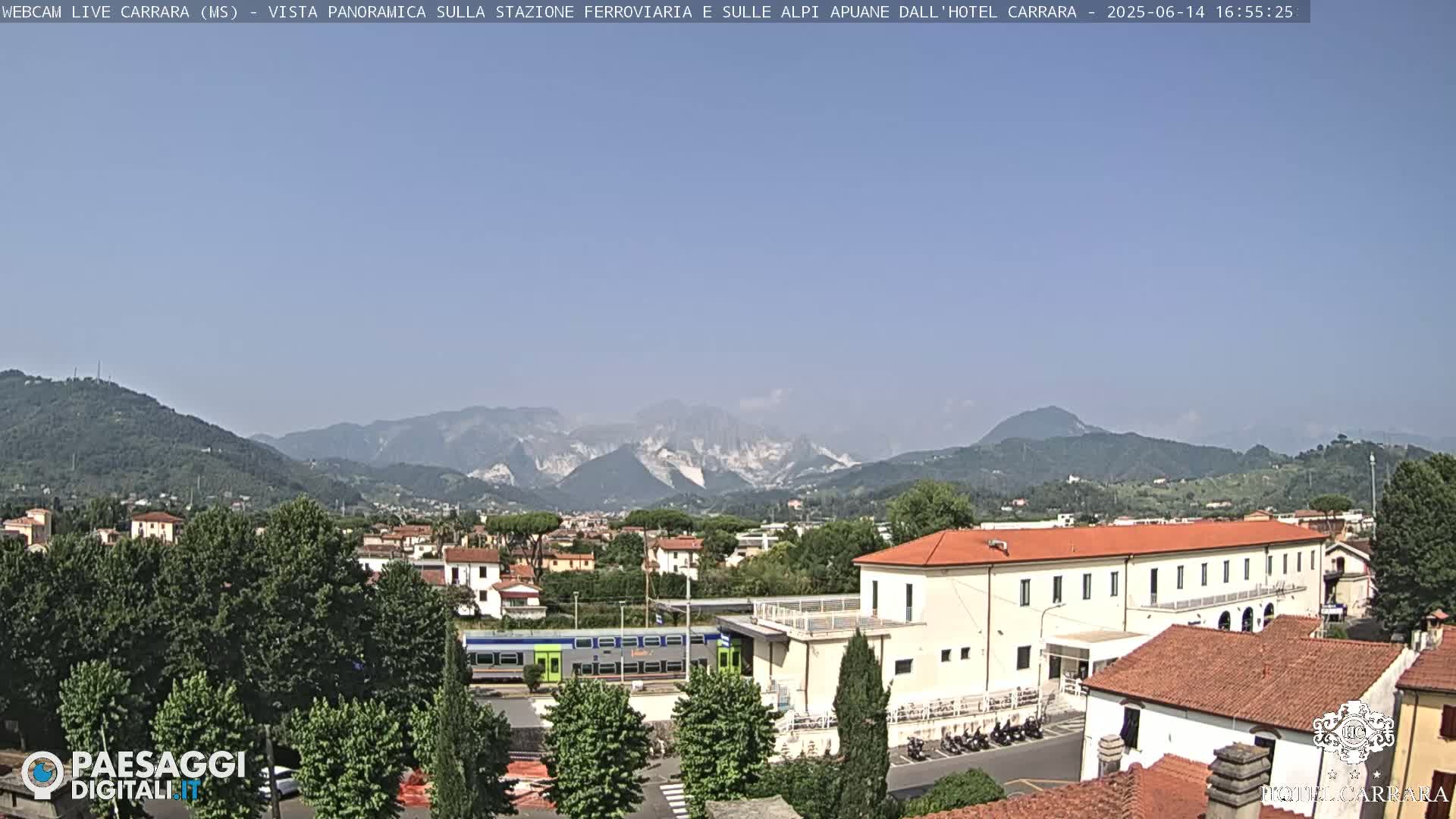 A long train sits in a station, surrounded by low buildings and lush green trees, with mountains in the background under a clear, sunny sky.