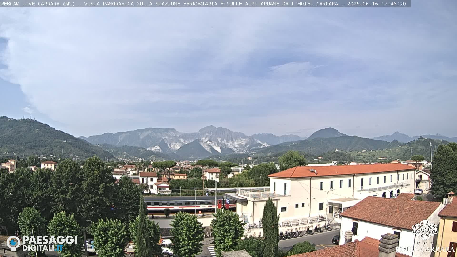 A partly cloudy day reveals a panoramic view of a train station nestled amongst buildings and lush green trees, with a mountain range in the background.