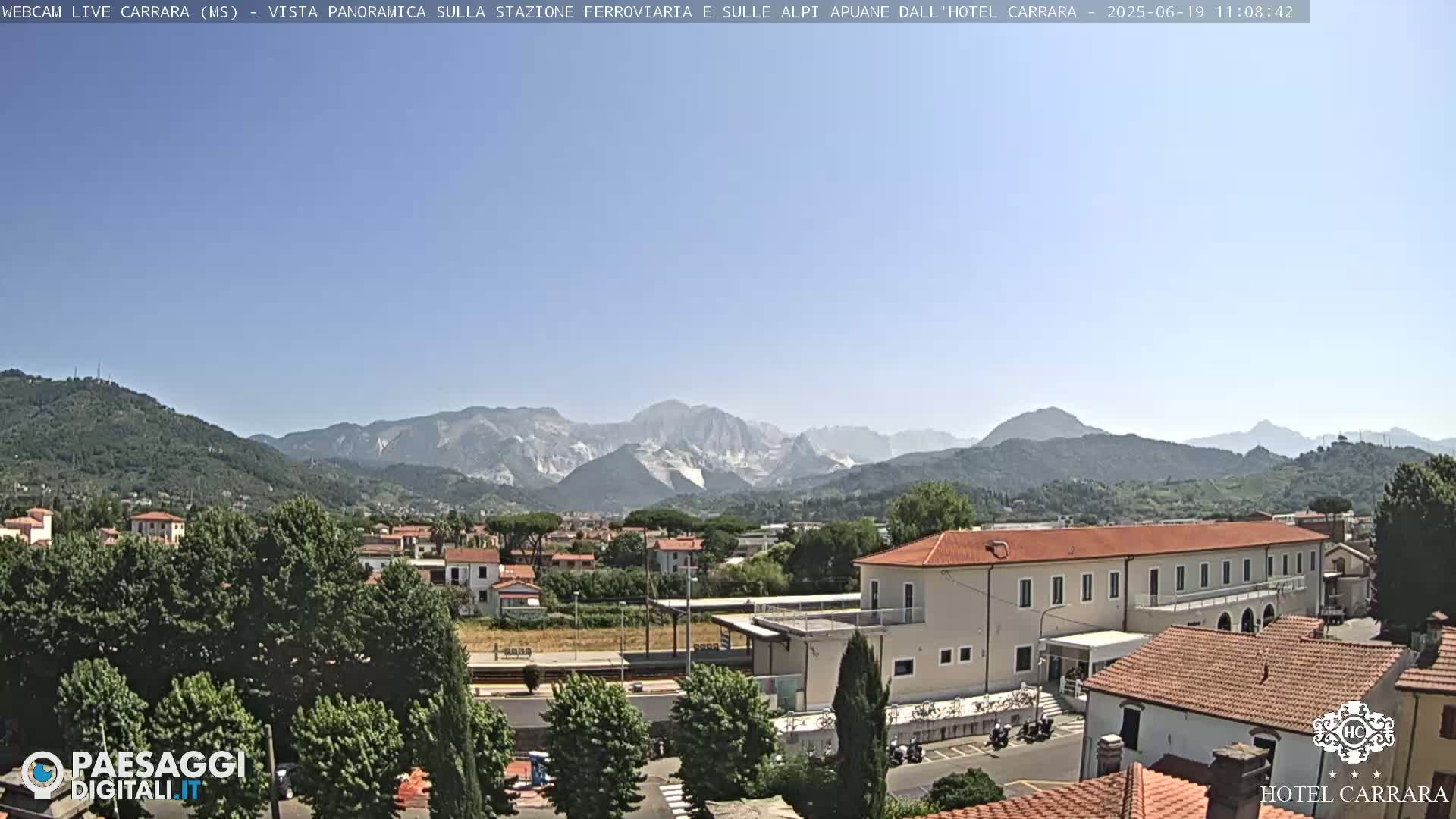 A panoramic view shows a town nestled in a valley, with mountains in the background under a clear blue sky.