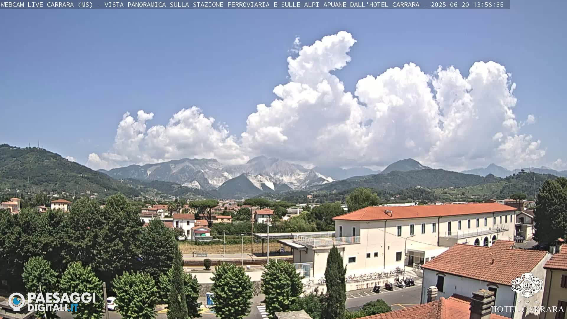 A partly cloudy day reveals a panoramic view of a town nestled in a valley, with mountains in the background and a building in the foreground.