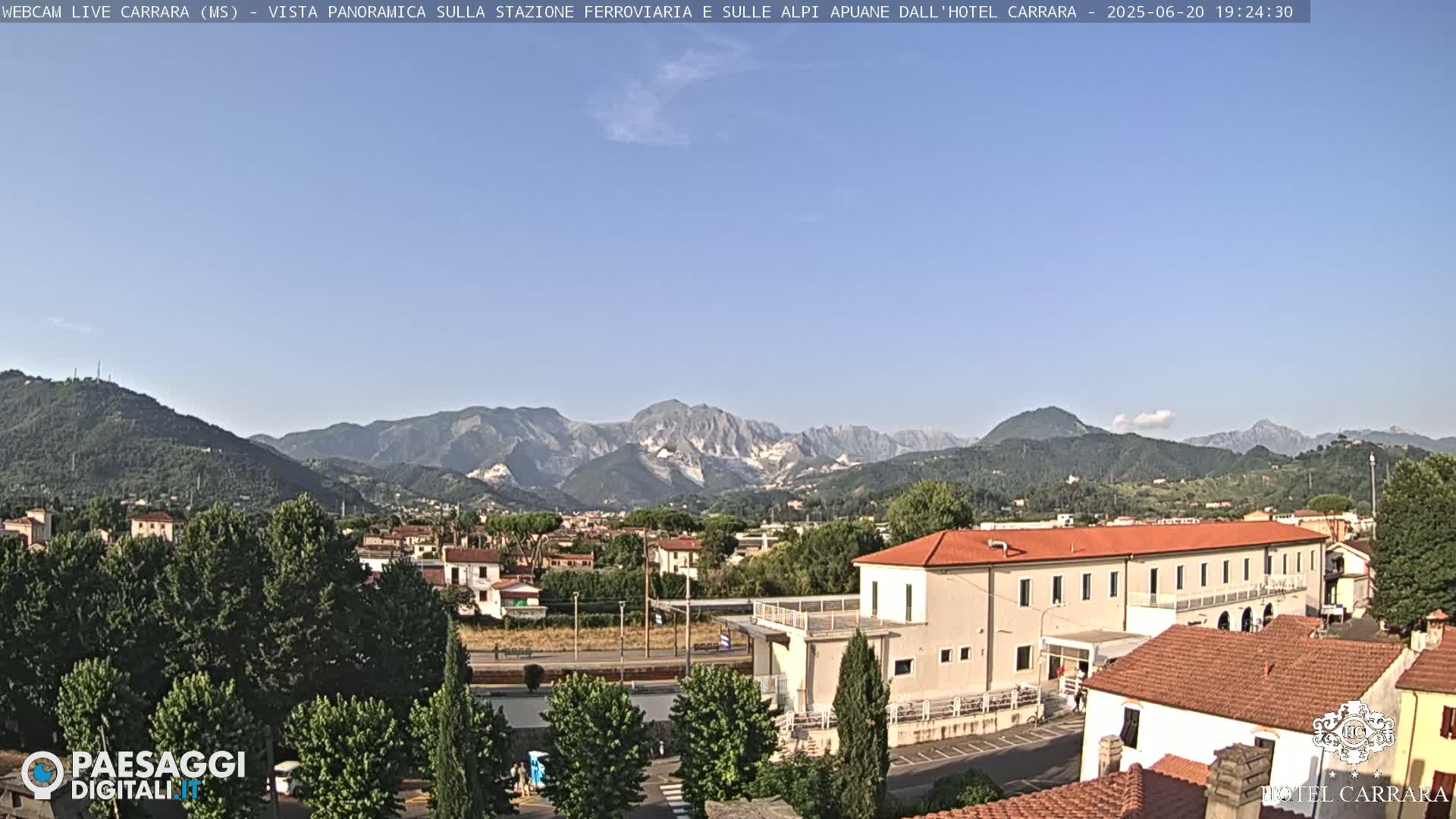 A panoramic view shows a town nestled in a valley, with a long, low building in the foreground and mountains in the background under a clear blue sky.