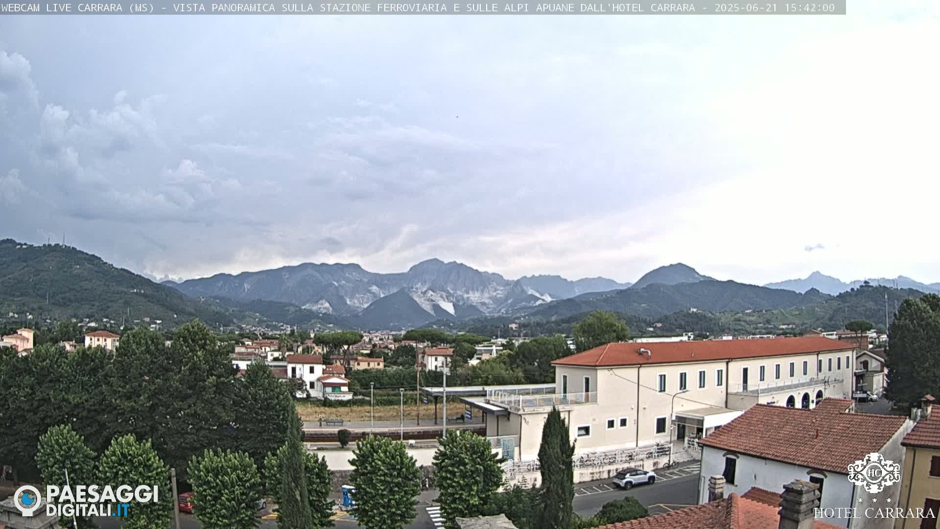 A partly cloudy day reveals a view of a town nestled in a valley, with mountains in the background and a long, low building in the foreground.