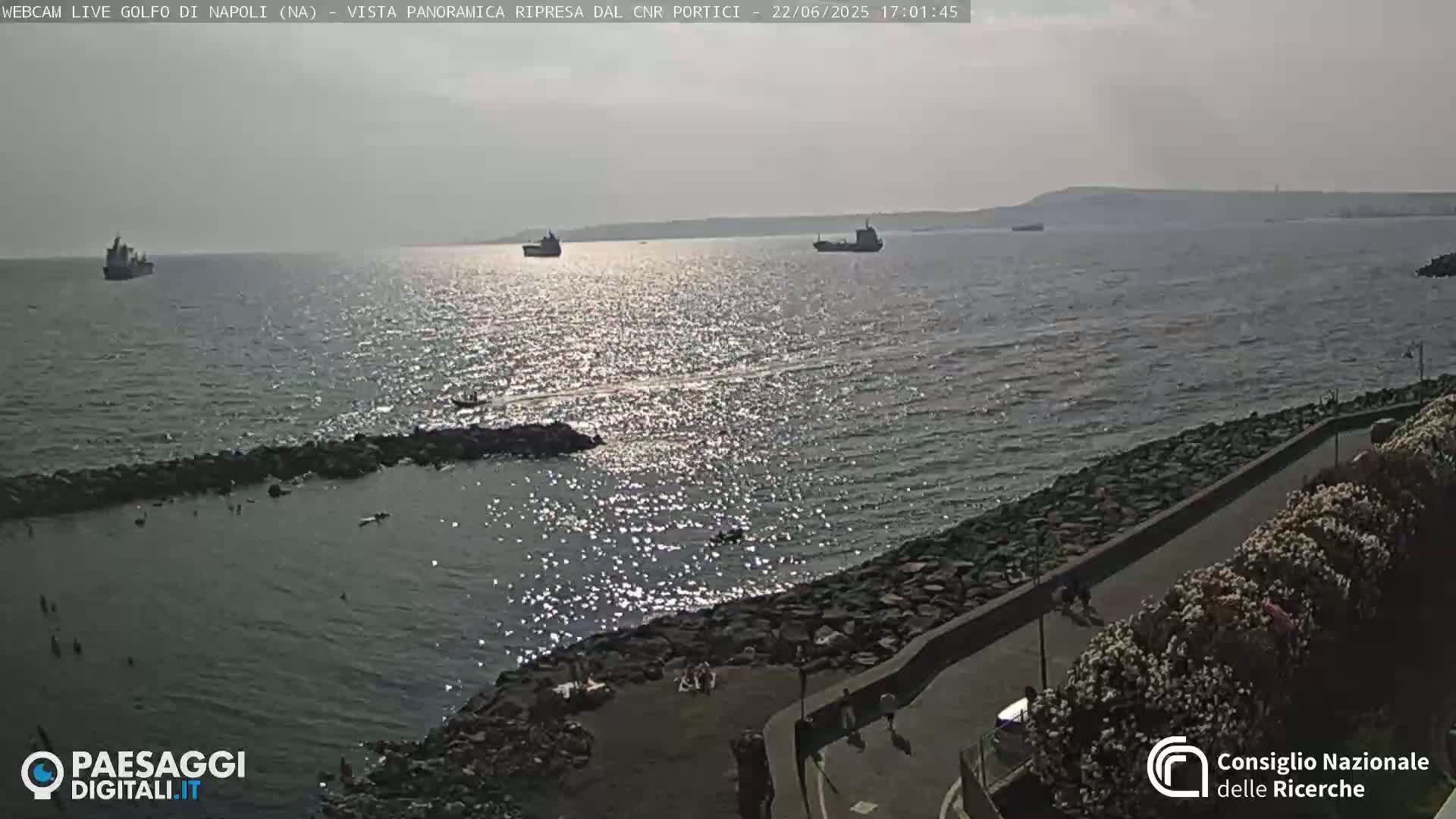 A sunny day shows a rocky shoreline with several people near the water, and a few ships visible in the distance on a calm sea.