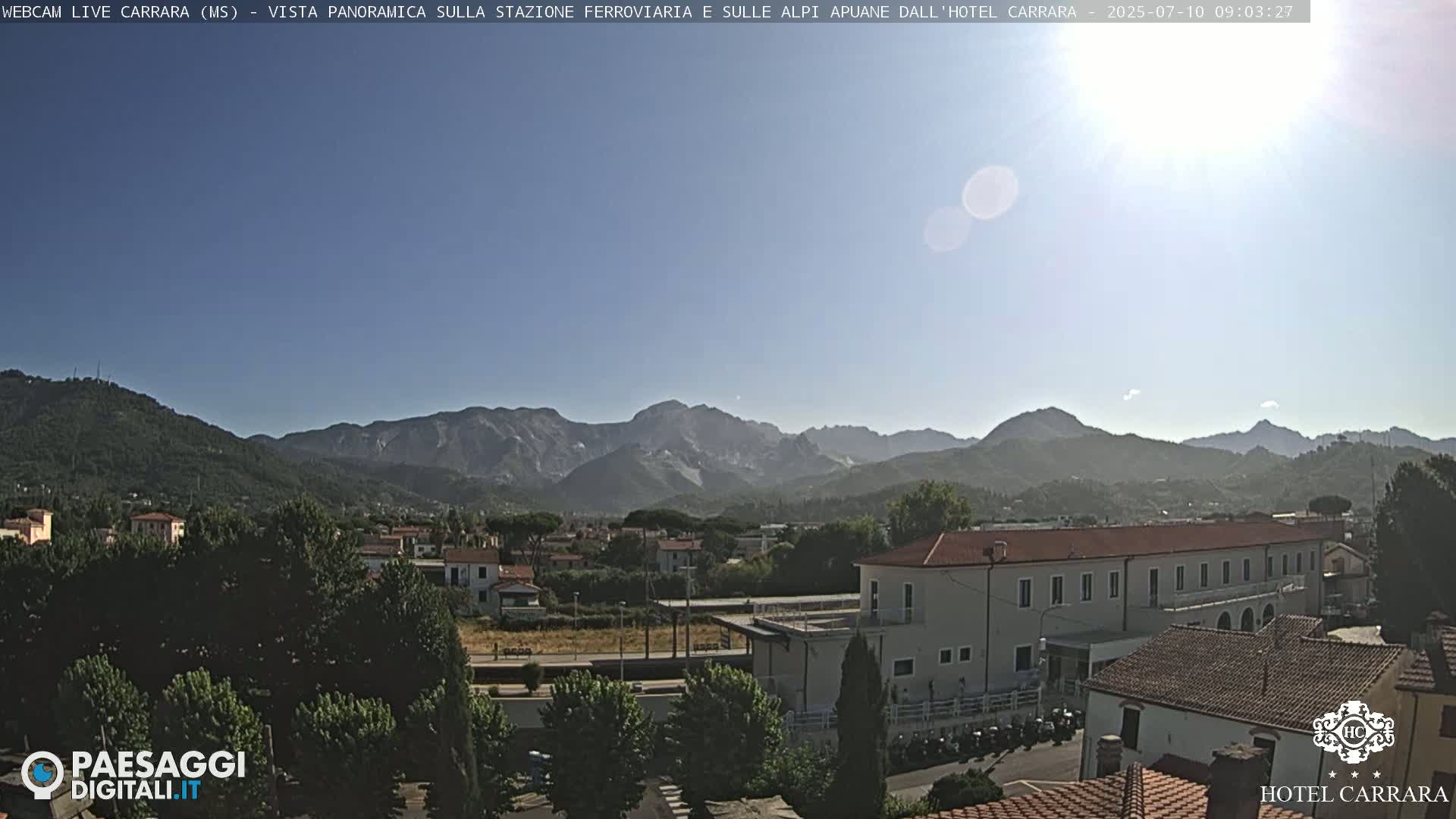 A sunny, panoramic view shows a town nestled in a valley, with mountains in the background.