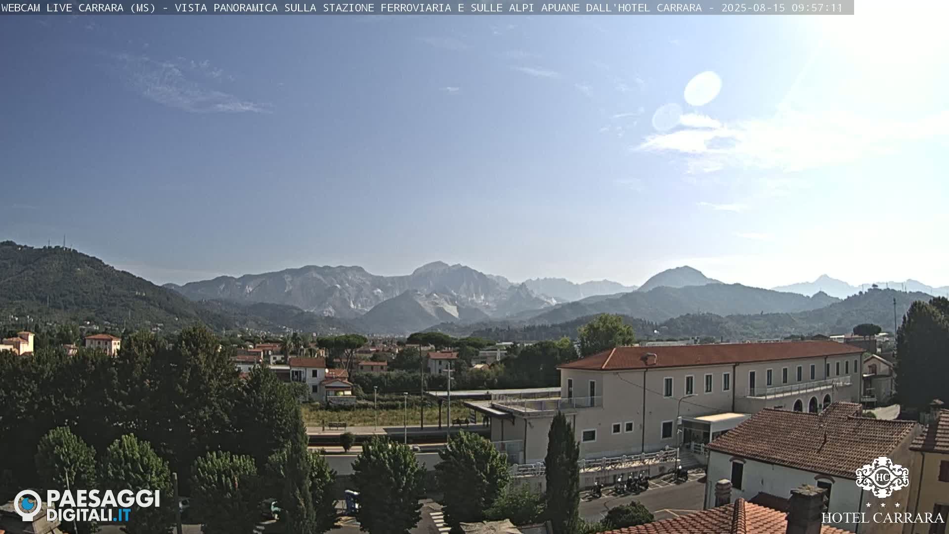 A mostly clear, sunny day shows a panoramic view of a town nestled in a valley, with mountains in the background.