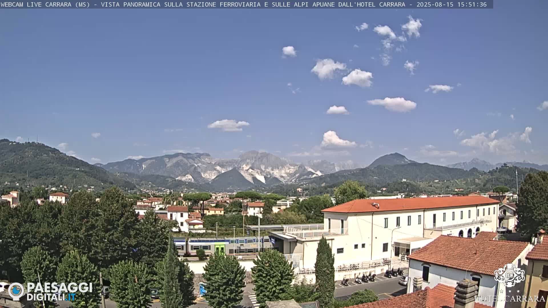 A mostly clear, sunny day shows a panoramic view of a town nestled in a valley, with mountains in the background.
