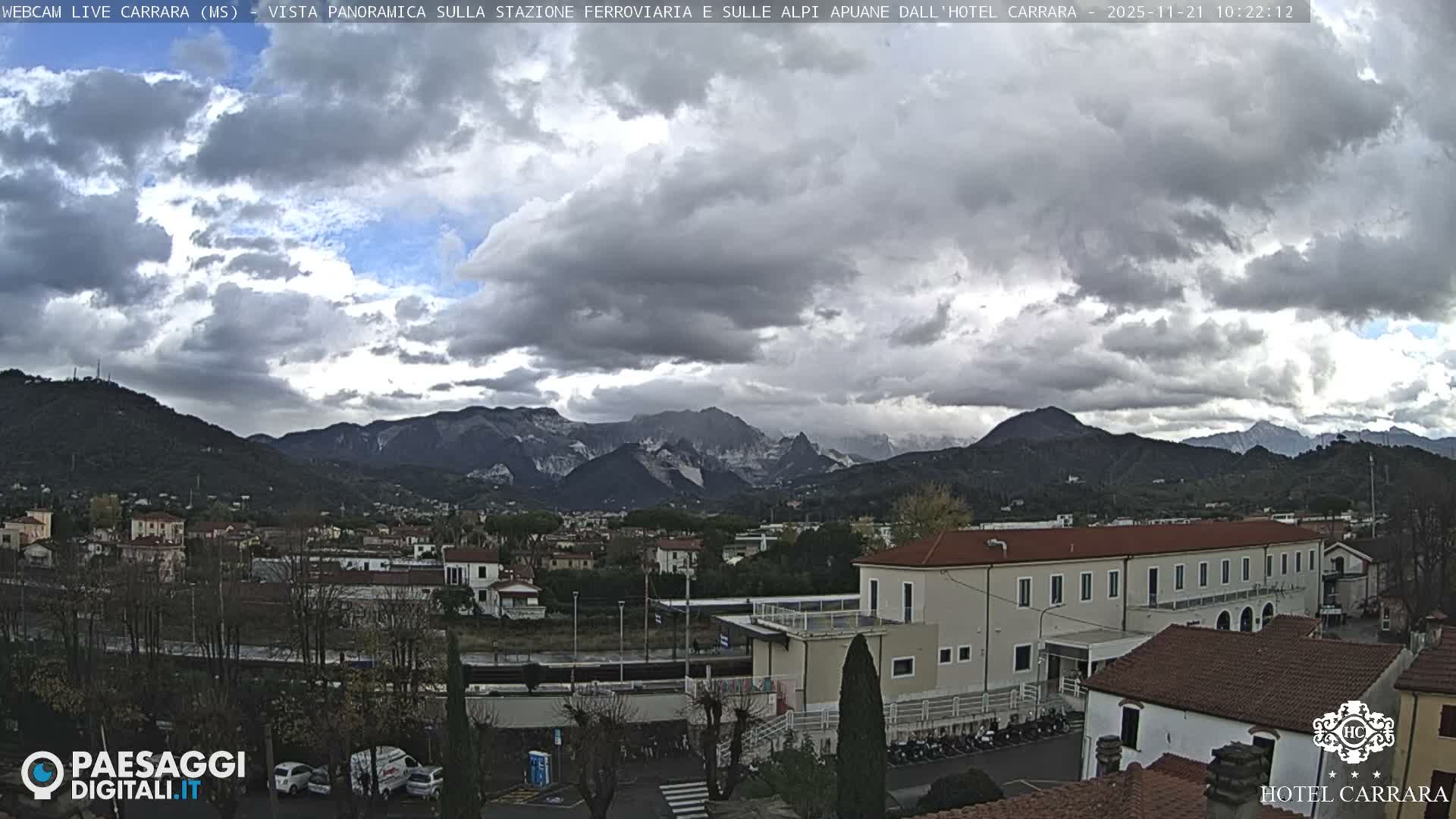 A panoramic view captures a town with buildings and sparse trees in the foreground, backed by imposing mountains featuring visible marble quarries, all under a predominantly overcast sky with patches of blue.
