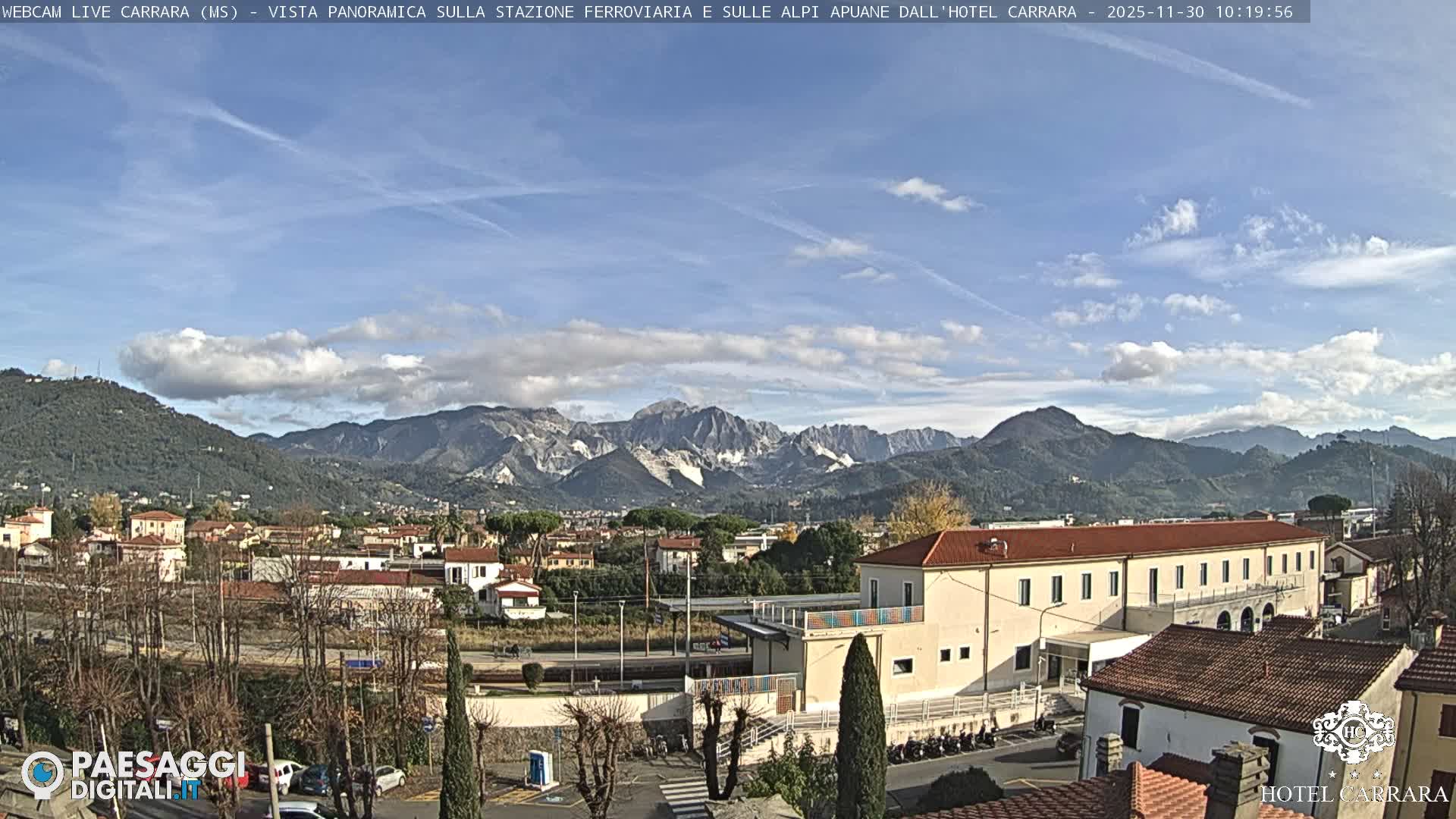 Under a clear blue sky dotted with scattered white clouds, a town with various buildings and a visible railway line sits in the foreground, backed by a majestic range of green, forested mountains, some revealing prominent white rock faces.