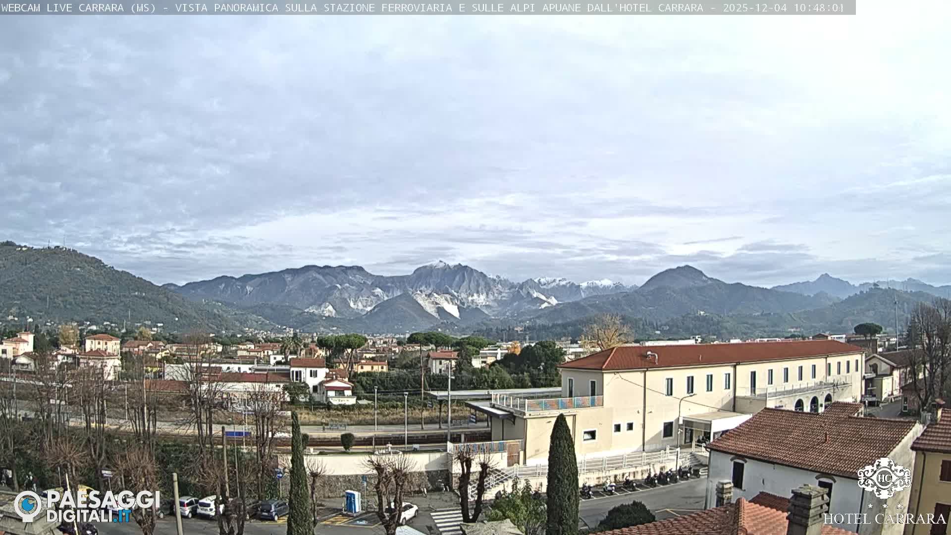 This panoramic image displays a town with various buildings and bare trees, set against a backdrop of imposing mountains featuring prominent white marble quarries and some distant snow-capped peaks, all under an overcast sky.