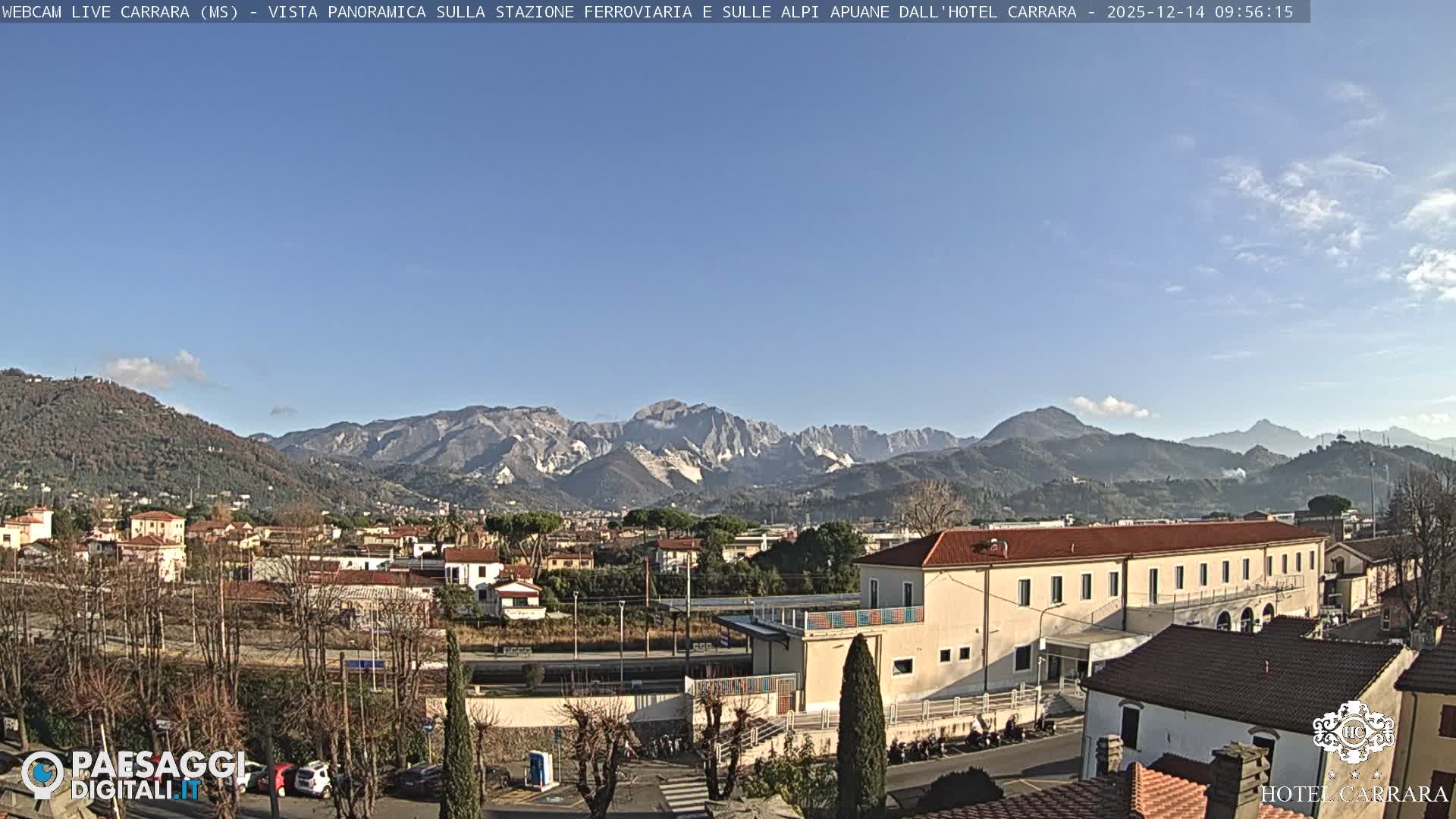 This panoramic image displays a town with various buildings and bare trees, set against a backdrop of imposing mountains featuring prominent white marble quarries and some distant snow-capped peaks, all under an overcast sky.