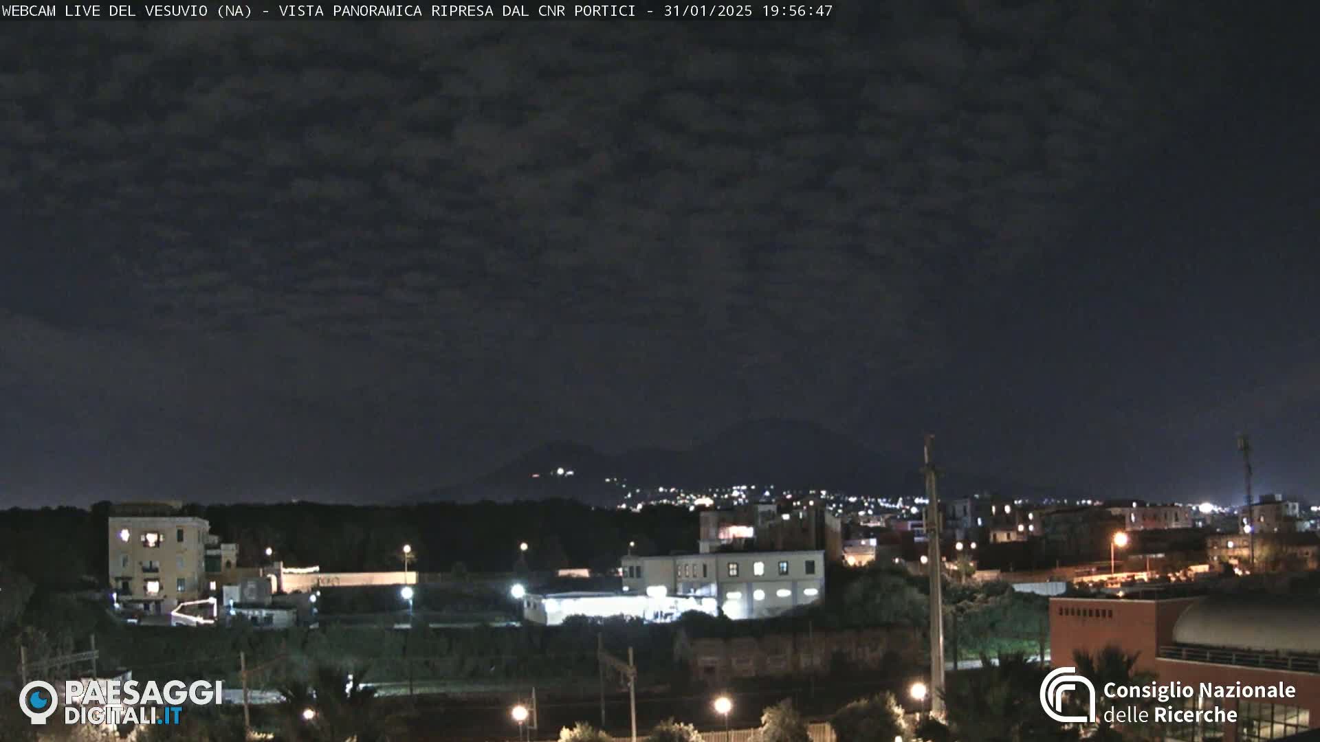 A nighttime view of a city with dimly lit buildings and distant mountains under a cloudy sky.
