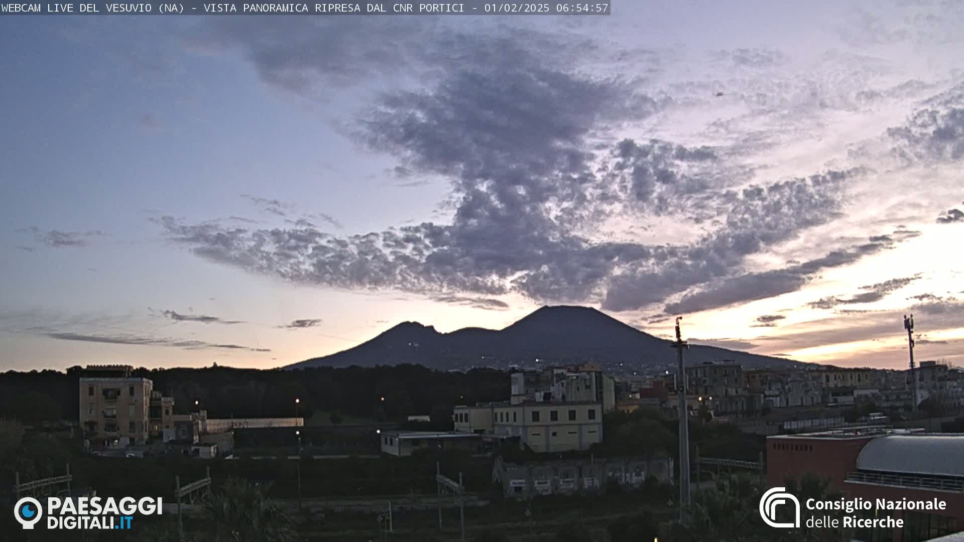A view of Mount Vesuvius at sunrise under a partly cloudy sky, with buildings and vegetation in the foreground.