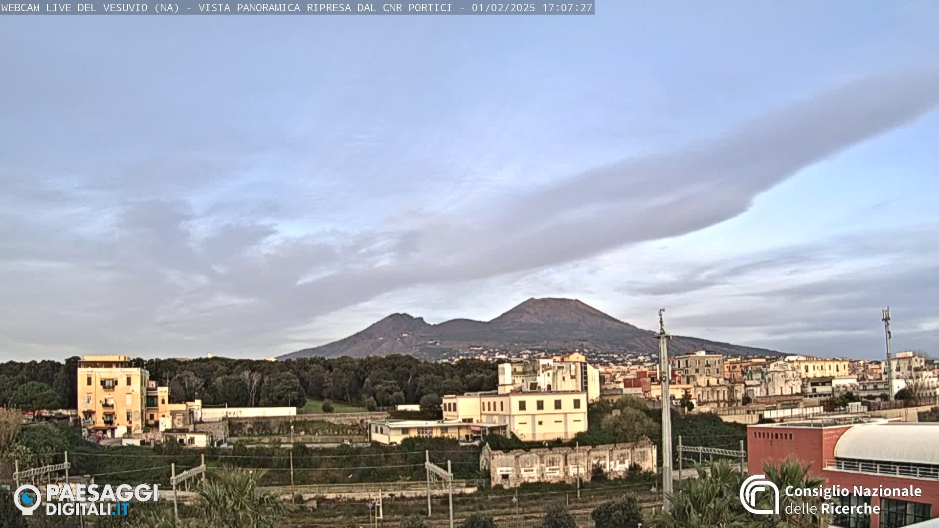 A view of Mount Vesuvius in the distance under a mostly clear, pale blue sky with thin, wispy clouds, overlooking a town with buildings and railway lines in the foreground.