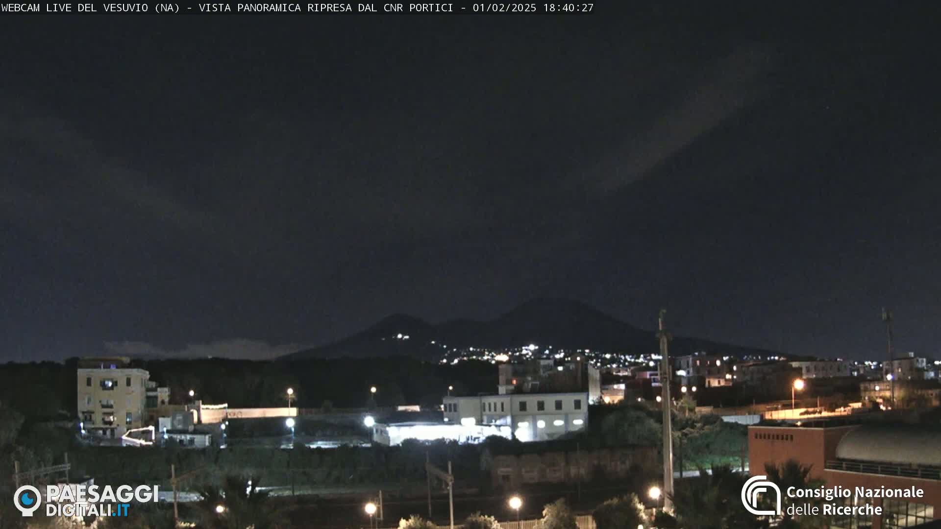 A nighttime view of a city illuminated by lights, with a dark mountain range visible in the background under a clear, dark sky.