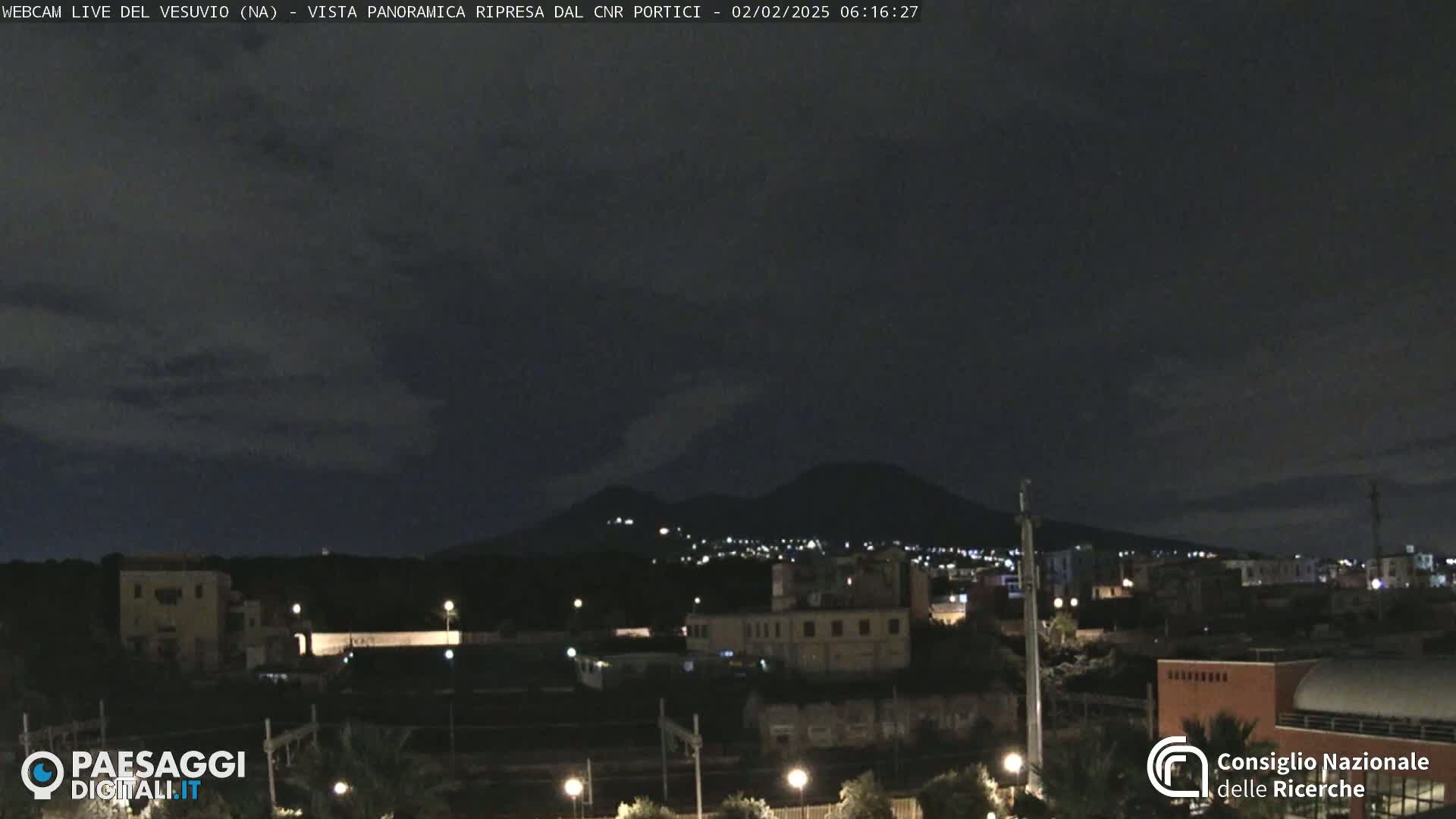A nighttime view of a city with a volcano in the distance under a partly cloudy sky.