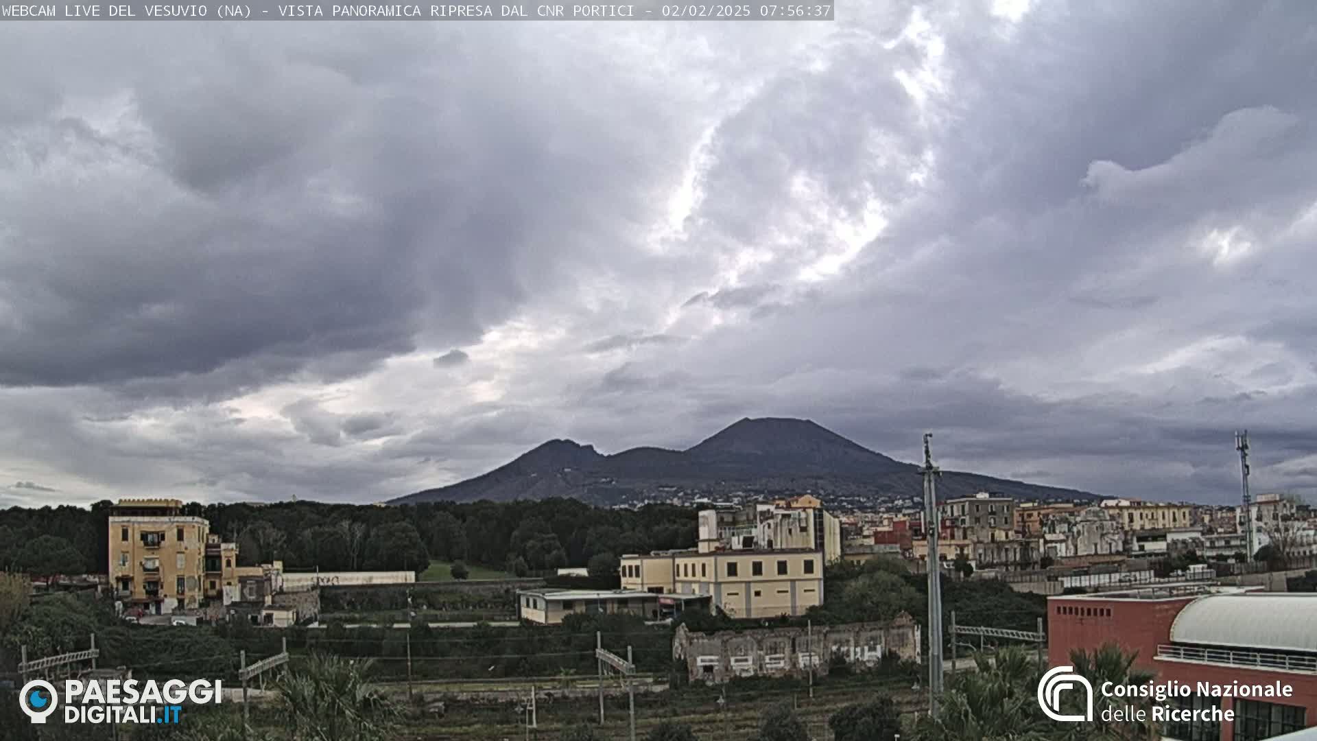 A view of Mount Vesuvius in the distance under a cloudy sky, with a town and buildings in the foreground.