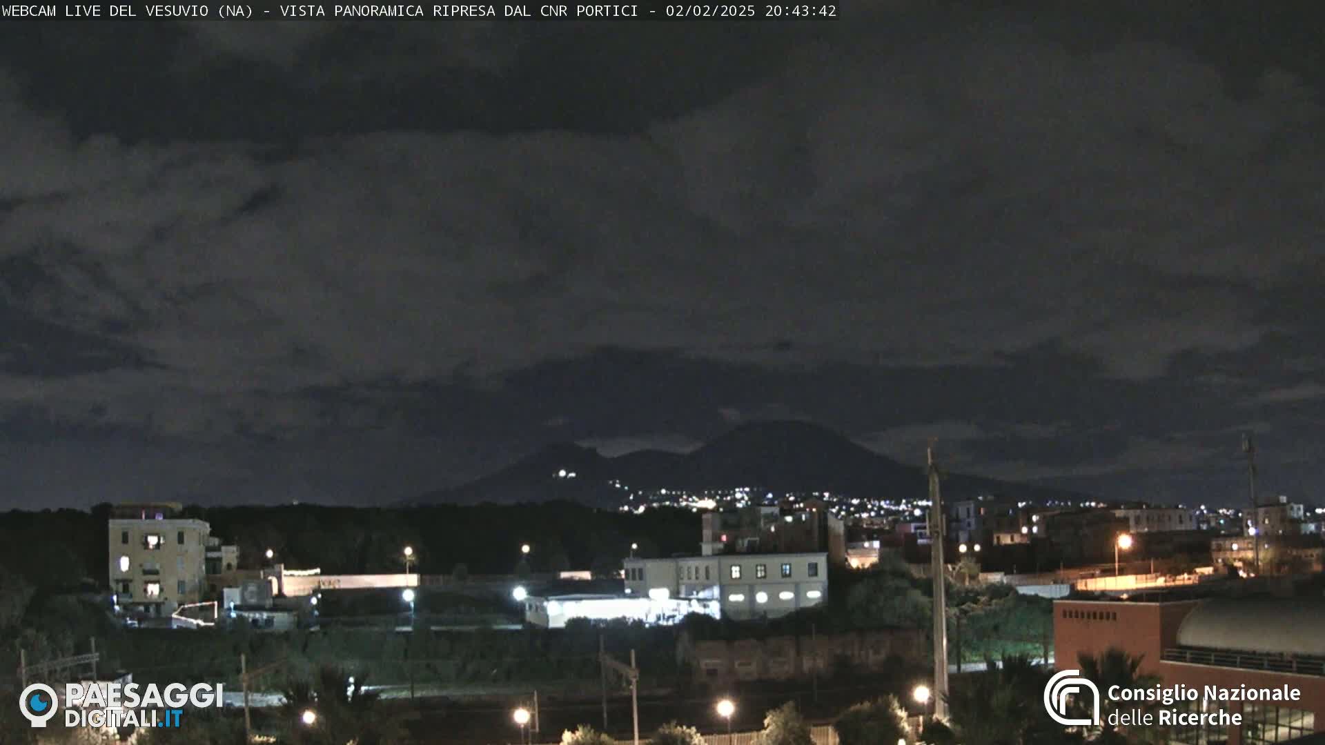 A nighttime view of a city illuminated with lights, with a dark, cloudy sky and a mountain range in the distance.