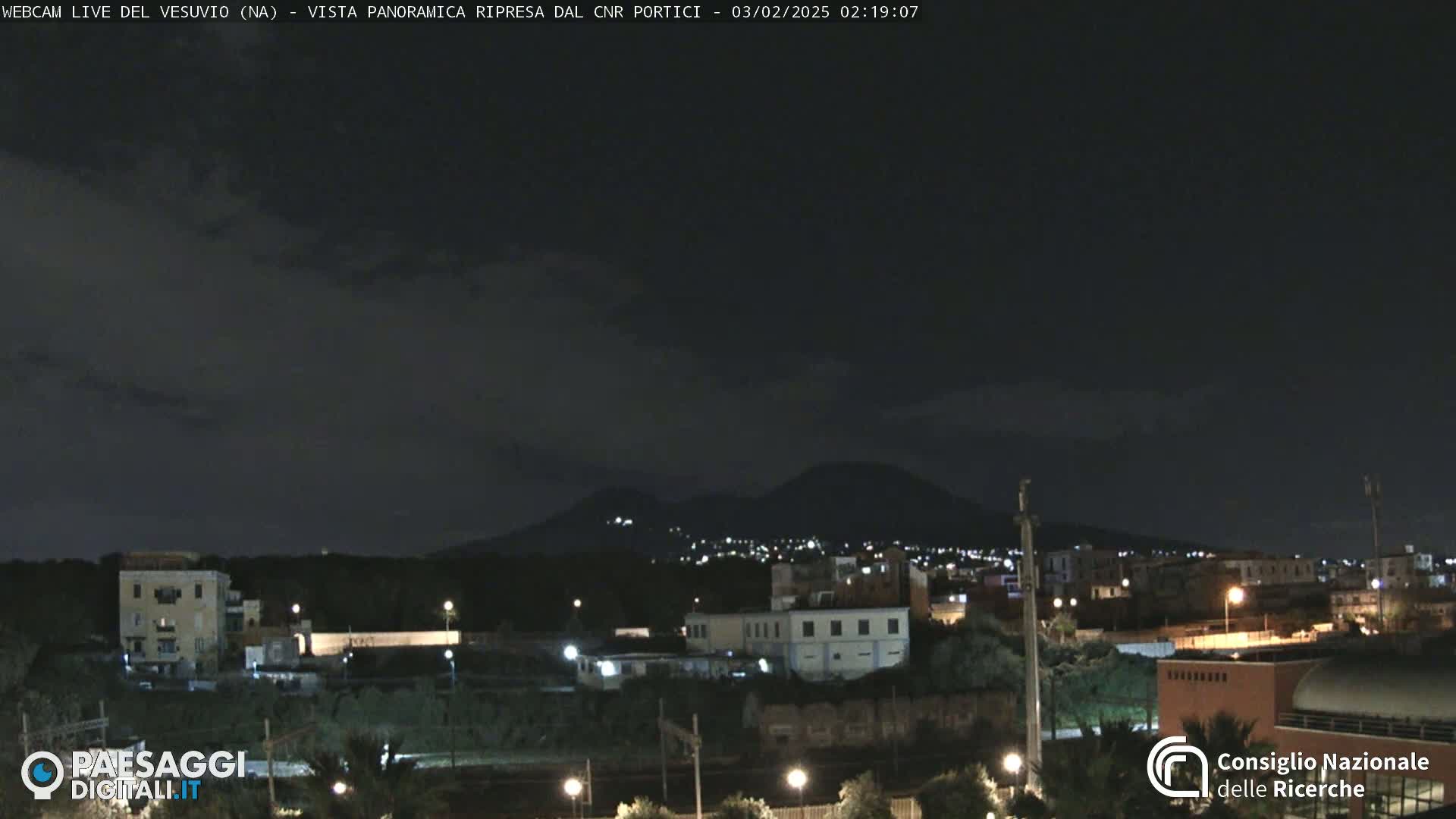 A nighttime view of a city illuminated by lights, with a dark mountain range in the background under a mostly clear sky.