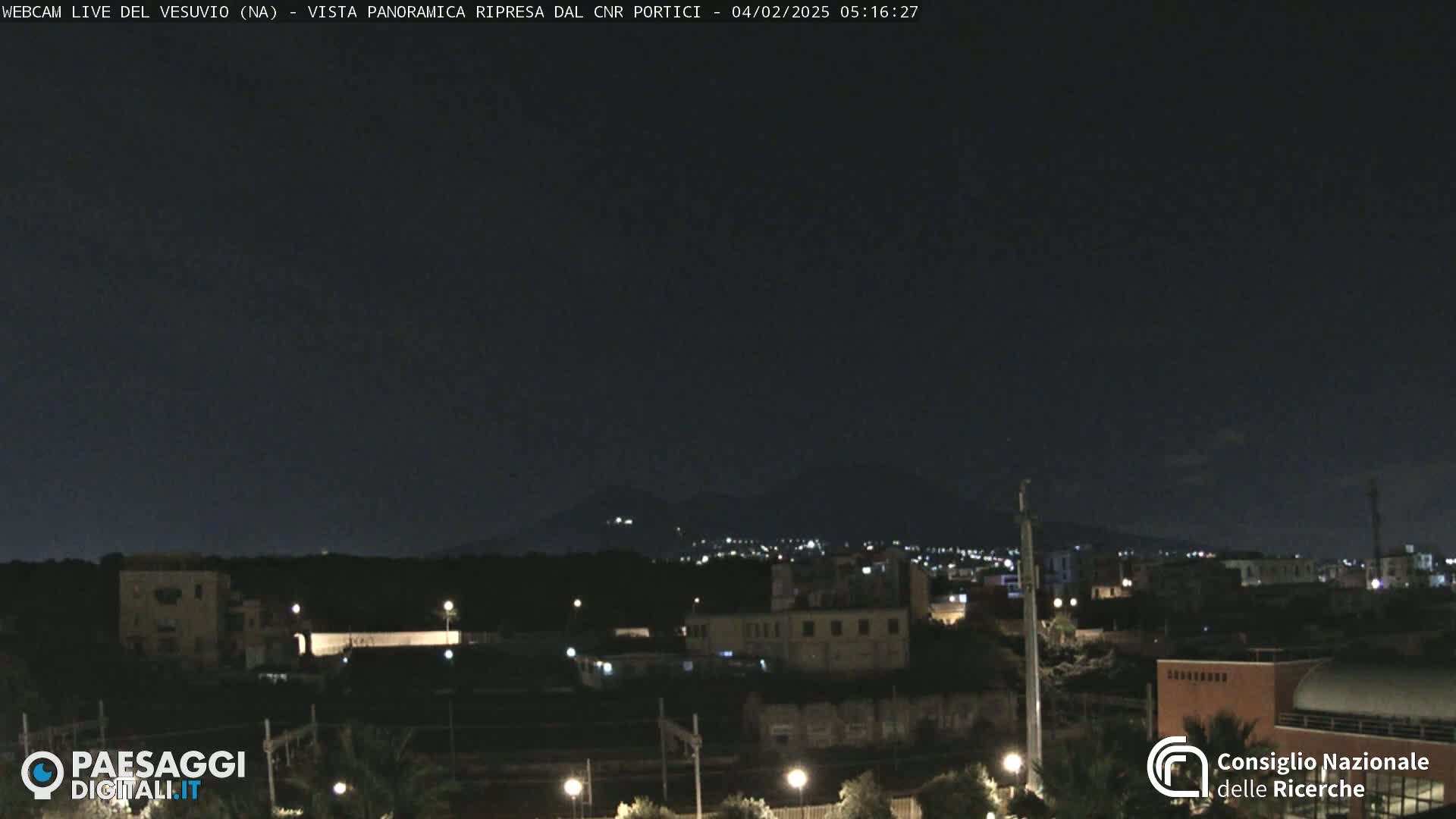 A nighttime, long shot of a city illuminated by lights, with a dark mountain range in the background under a clear, dark sky.