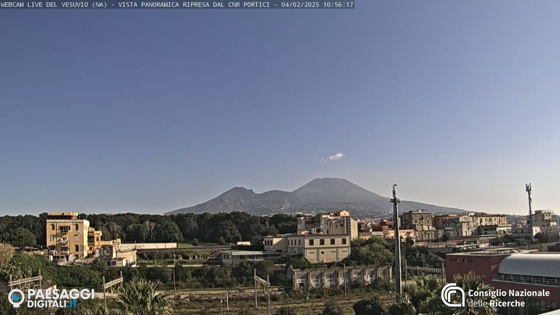 A clear, sunny day reveals Mount Vesuvius in the distance, overlooking a town with buildings and greenery in the foreground.