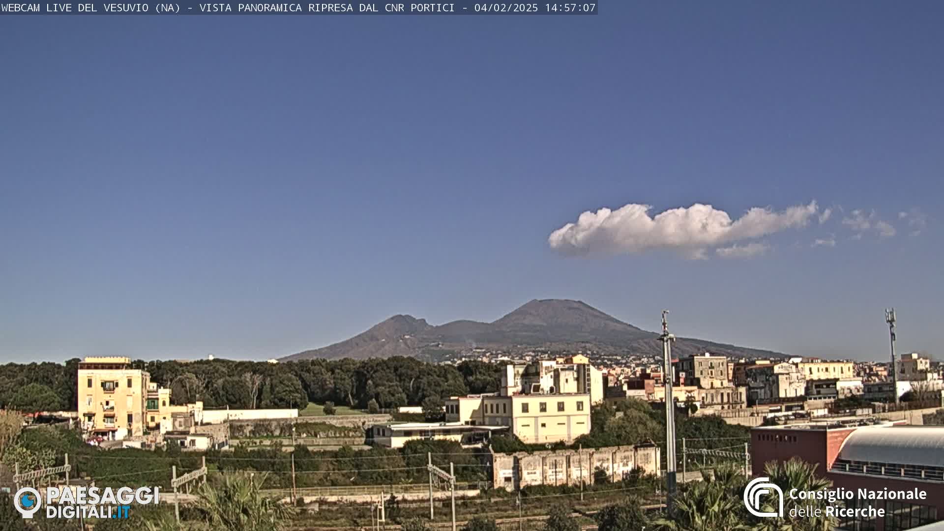 A view of Mount Vesuvius in the distance, under a mostly clear blue sky with a single, puffy cloud, shows a town with low buildings and trees in the foreground.