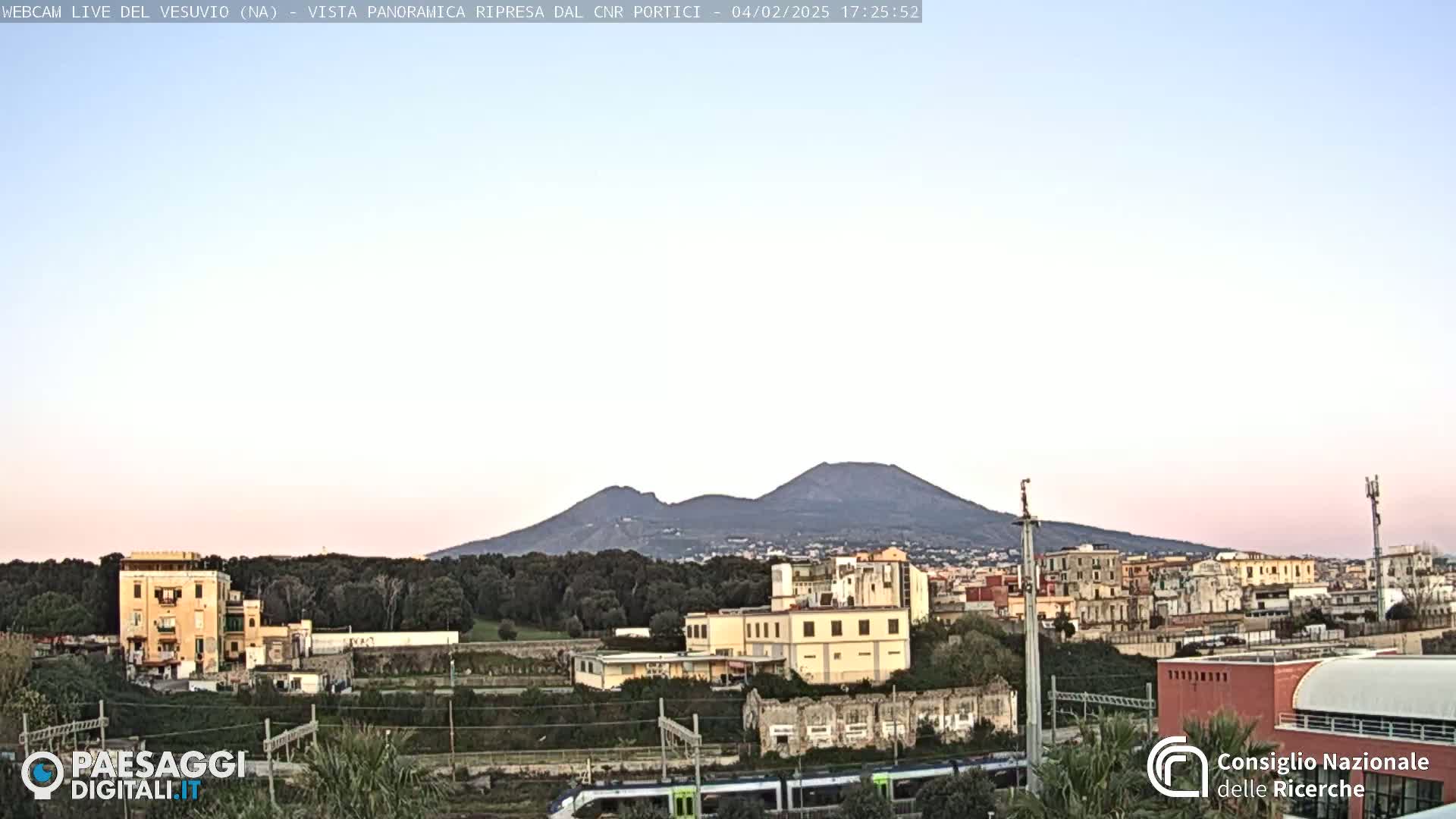 A distant view of Mount Vesuvius under a pale pink and light blue sky shows a cityscape and train tracks in the foreground.