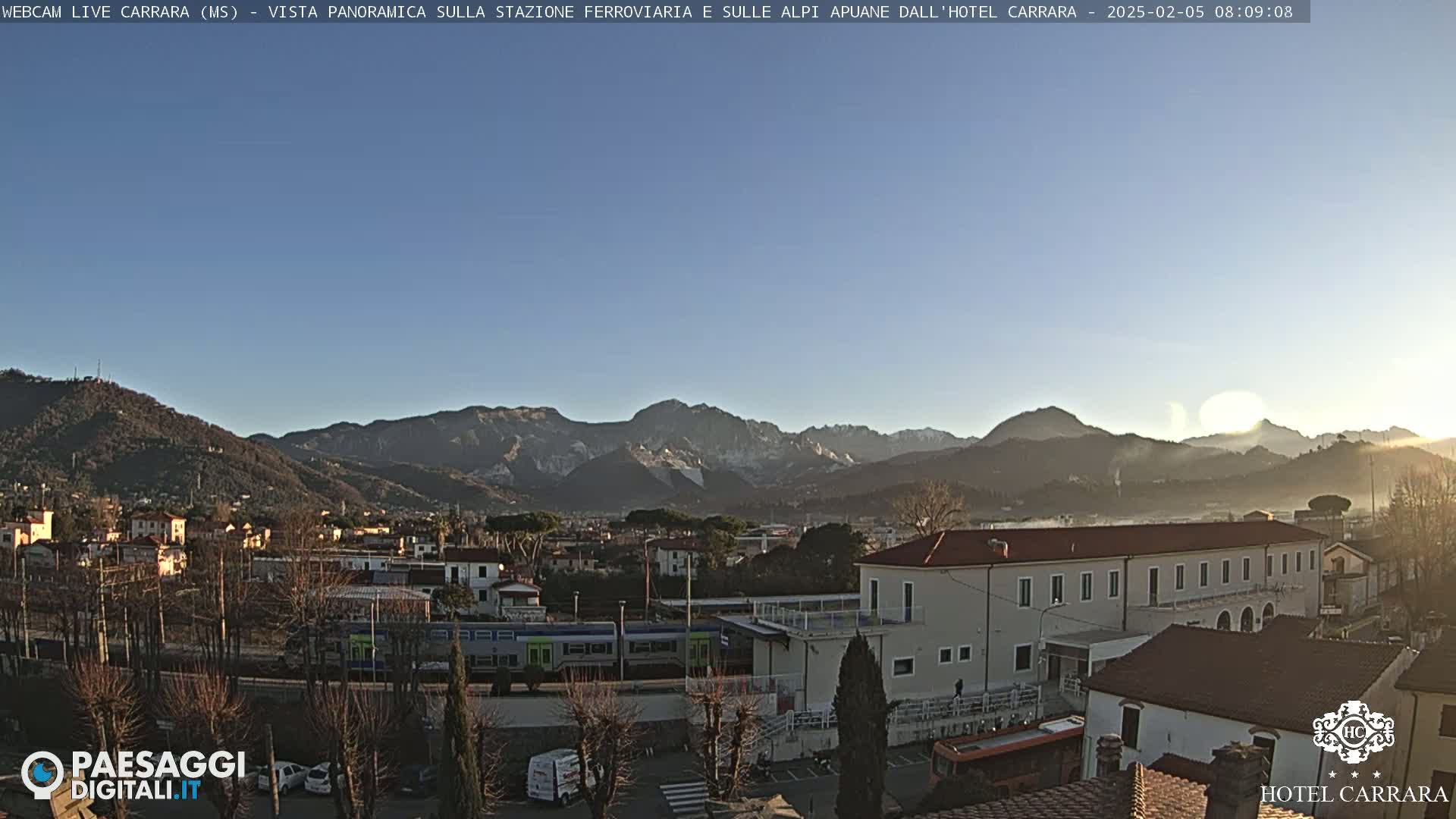 A clear, sunny morning view of a town nestled in a valley, with mountains in the background and a train visible near a building.